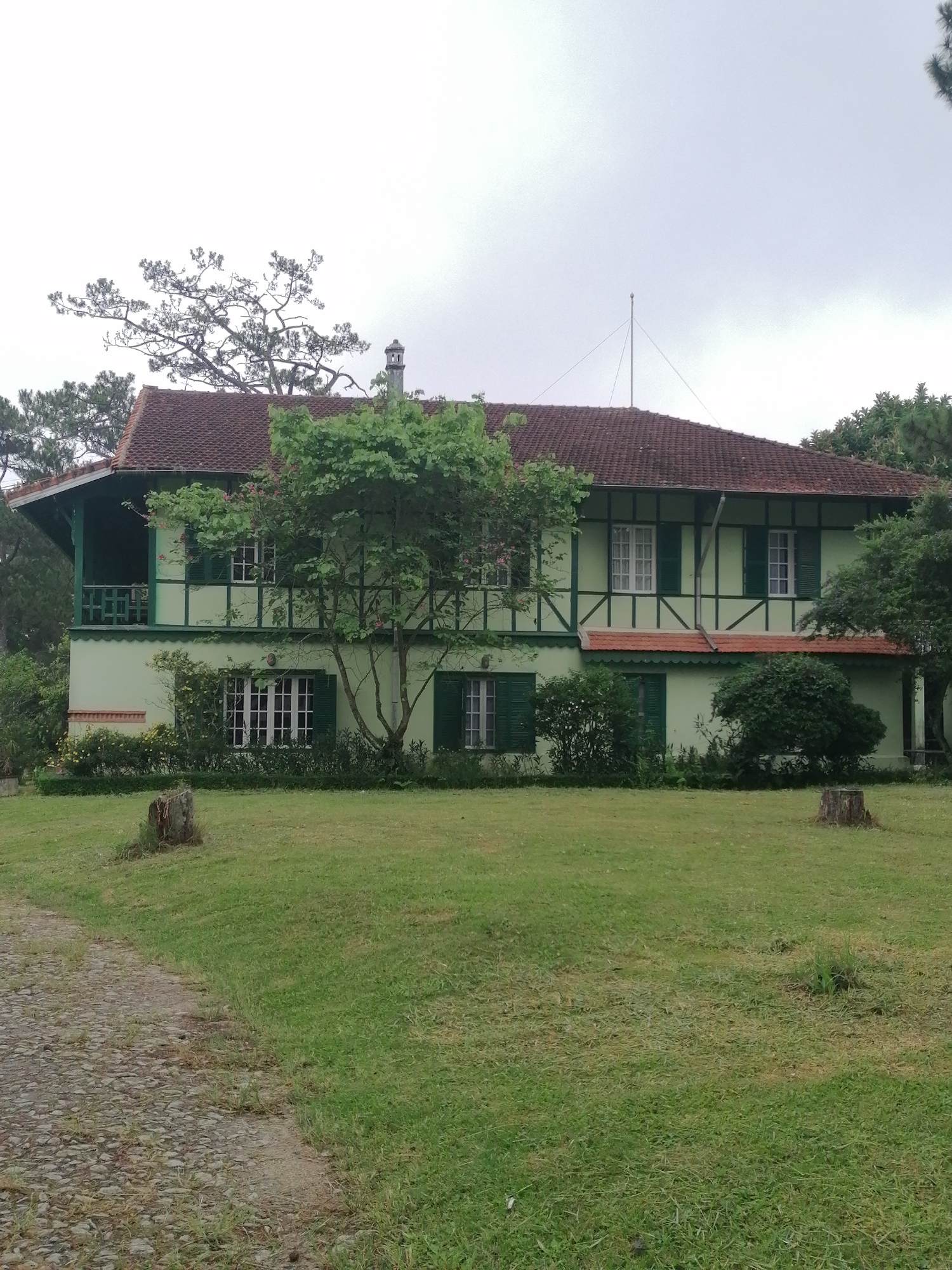 The image captures a serene, two-story house nestled amidst lush greenery under an overcast sky. The house, possibly of colonial or European influence, features light green walls with striking dark green trim, including decorative half-timbering on the upper floor. Its roof is made of dark red or terracotta tiles, with a steep pitch.
On the left side of the house, there's a covered balcony with a dark green railing, and a chimney is visible rising from the roof nearby. Another smaller, decorative balcony or railing is present on the upper right side of the facade. The windows are framed in white, many adorned with dark green shutters, adding to the house's quaint charm. An antenna or mast extends from the roof towards the right side.
The house is surrounded by abundant vegetation. Several trees and shrubs are situated directly in front of and around the building, with a particularly prominent tree standing in the foreground, partially obscuring the house. More trees and dense foliage form a backdrop, hinting at a natural, secluded setting.
In the immediate foreground, a vast, neatly mown green lawn stretches out, punctuated by two rough tree stumps. A cobblestone path or driveway is visible on the far left, leading towards the house. The sky is uniformly gray and overcast, suggesting a cloudy day without direct sunlight, contributing to a calm and somewhat muted atmosphere. There are no people visible, and no specific activity or event is taking place, presenting a tranquil, static scene. The architecture and verdant surroundings are consistent with the location context of Da Lat, Vietnam. No visible text is present in the image.