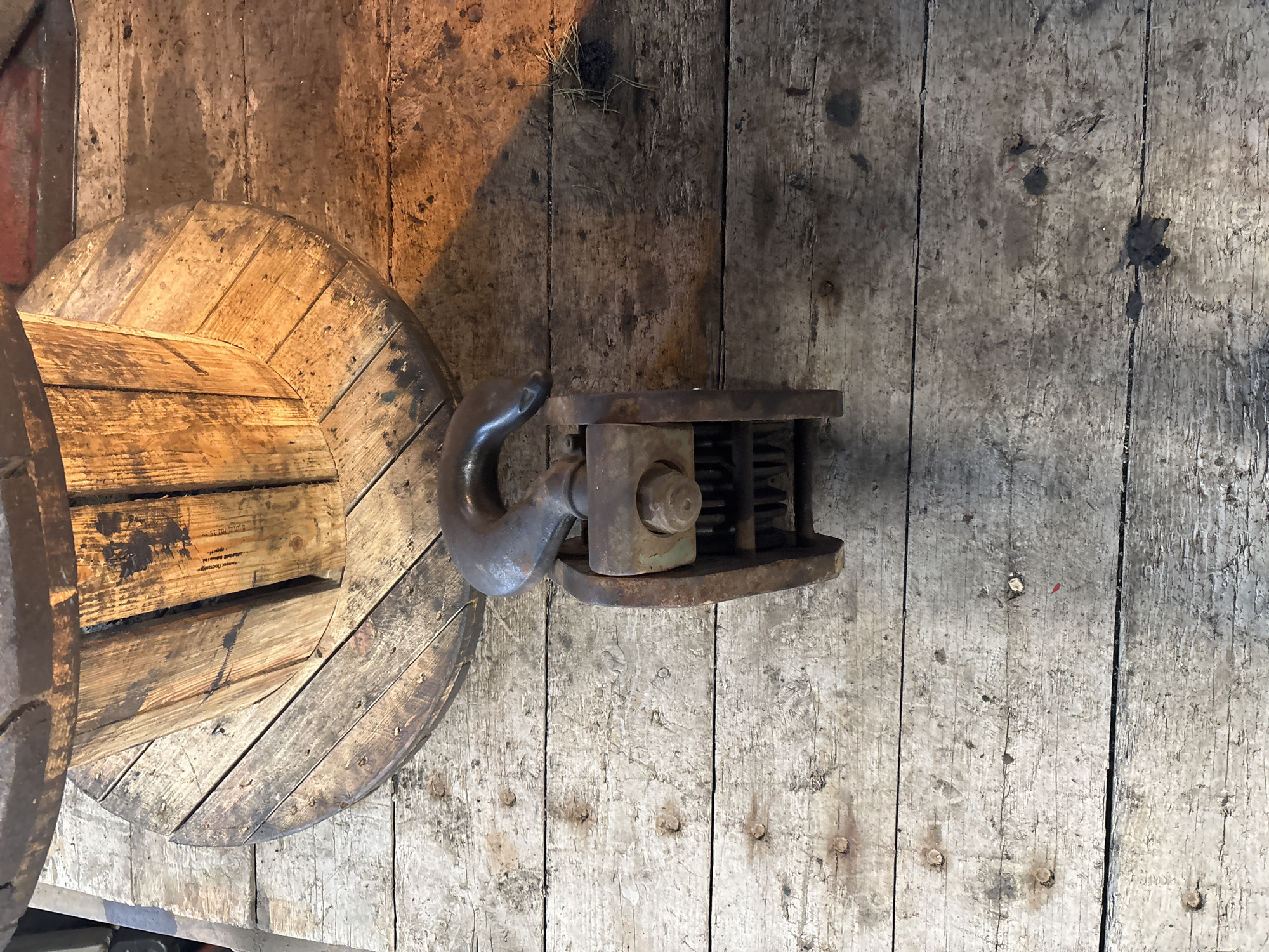 The image displays an overhead shot of a large, weathered wooden spool and a rusty metal pulley system resting on a rustic wooden floor. The spool, made of planks of wood, is worn and appears to have been used for storing heavy cables or ropes. The pulley system, made of dark, oxidized metal, features a large hook and a series of wheels, suggesting it's designed for lifting or moving heavy loads.
The wooden floorboards are aged and marked with dirt and scuffs, indicating an industrial or storage setting. A shadow falls across the floor from the top left, casting a diagonal line and hinting at a light source, possibly a window or overhead lamp. There is a small clump of dried grass or straw on the wooden surface near the top edge of the image.
The scene conveys a sense of history and utility, with the aged materials suggesting a long period of use. The lighting creates a moody atmosphere, highlighting the textures and imperfections of the objects and their surroundings. There are no people visible in the image. There is some faint text visible on one of the wooden planks of the spool, but it is too indistinct to be read. The location context of Saint Petersburg, Russia, doesn't offer specific visual cues within the image itself.