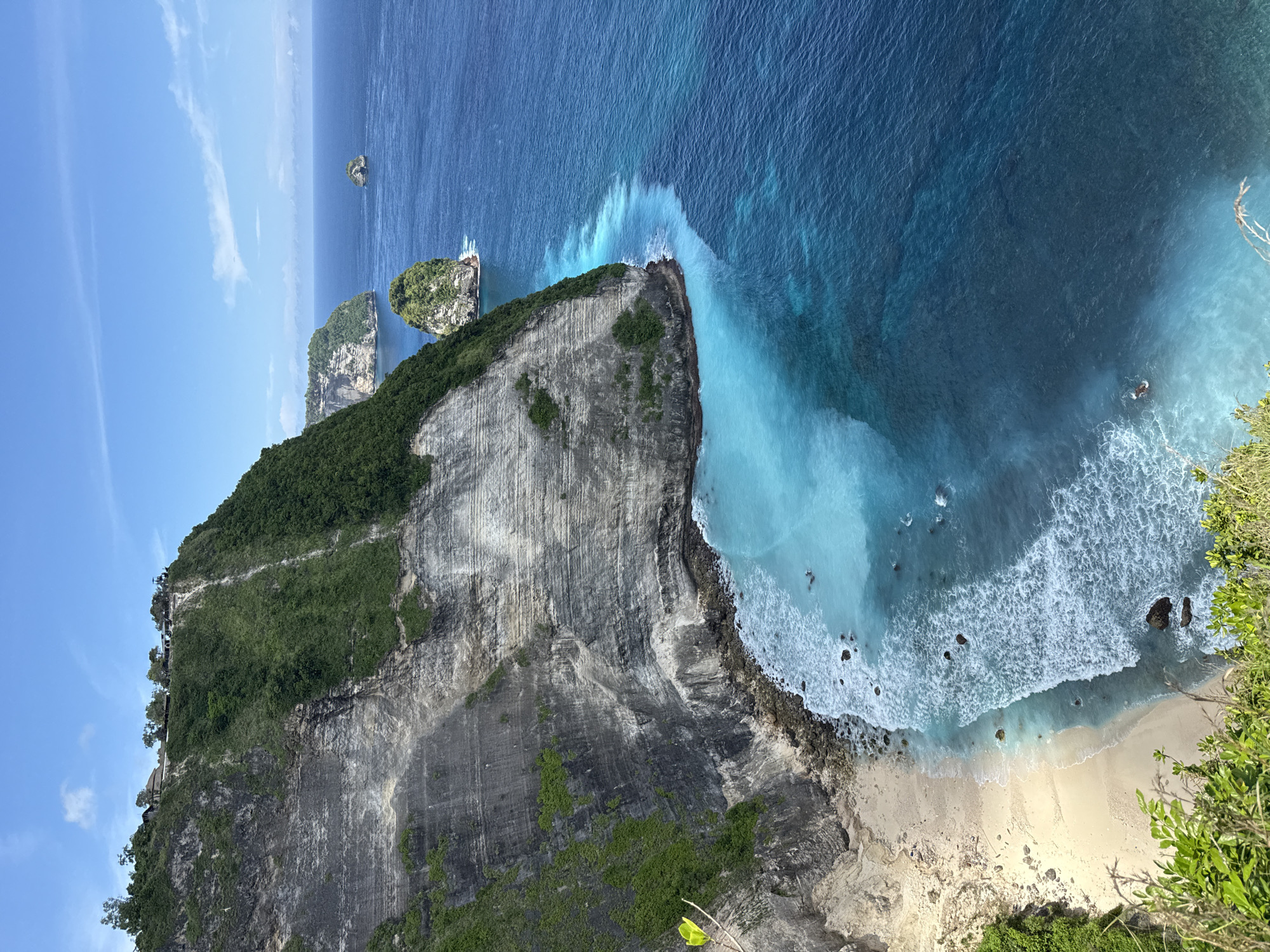 The image shows a coastal scene with a massive cliff face dominating the view. The cliff is layered with grey rock and some vegetation, mainly green shrubs. Below the cliff, there is a small sandy beach with visible waves crashing onto the shore. The water transitions from a deep blue color further out to a light turquoise near the shore, where the wave action is most visible. In the distance, there are a couple of smaller rocky islands with vegetation. The sky is blue with some scattered clouds.
This appears to be a view of Kelingking Beach, located in Bunga Mekar, Indonesia. The steep, rocky cliff face and the characteristic shape of the beach are consistent with this location.