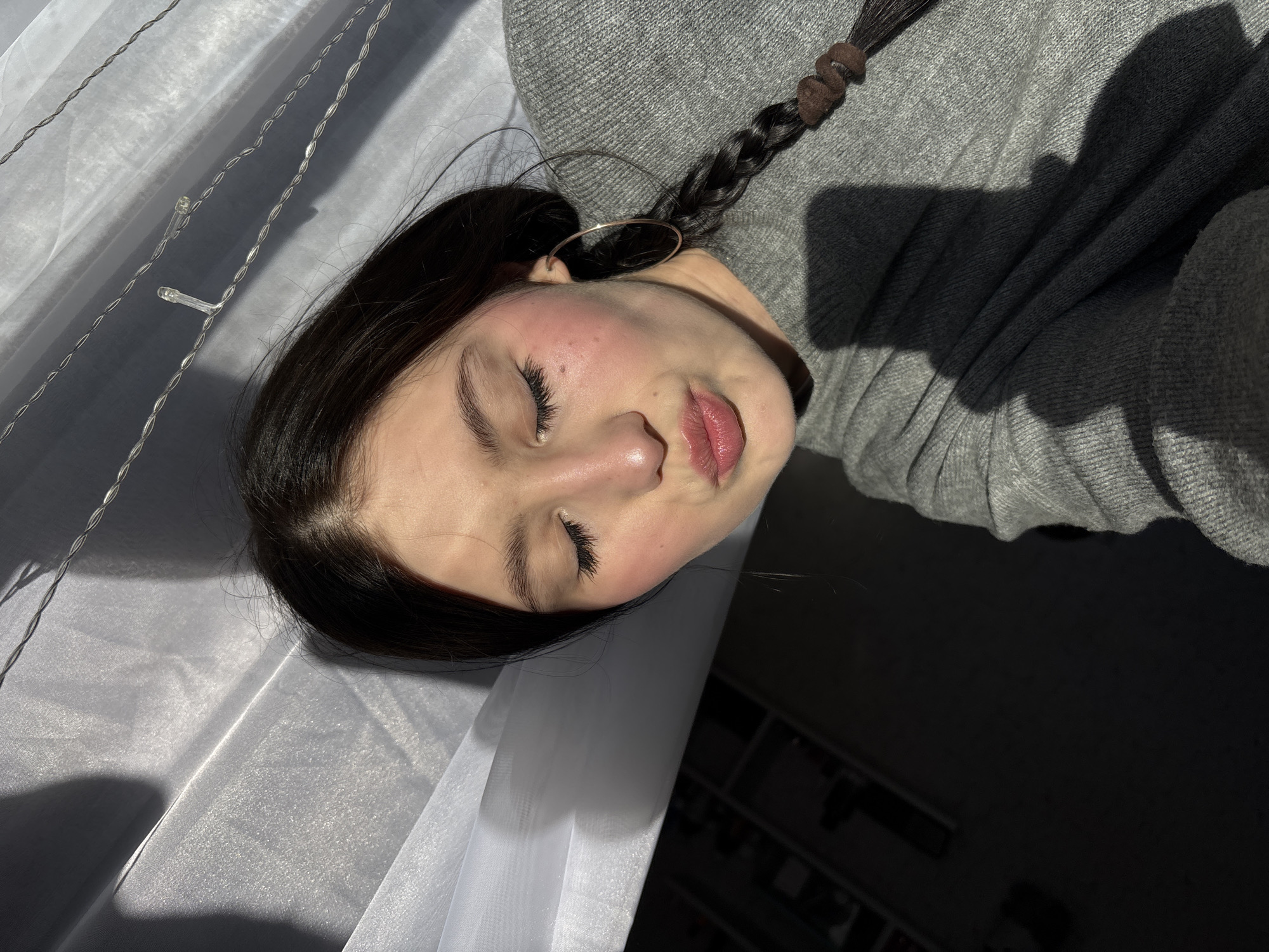 A young woman with dark hair, styled in a braid, lies on a white surface. She wears a gray sweater and a hoop earring. Her eyes are closed. Part of the scene is in shadow, possibly due to natural light. There is no indication of the location being Фоки, Russia.