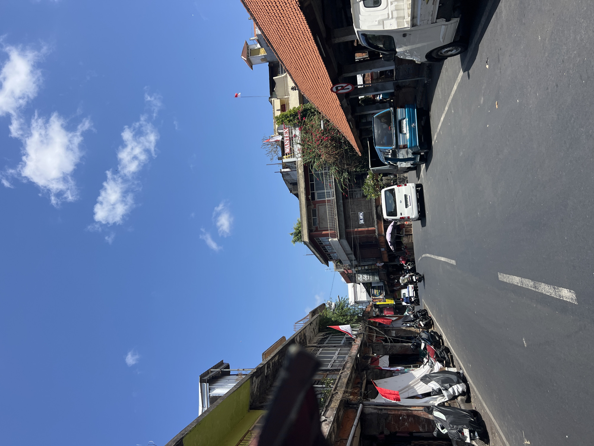 This is a daylight, eye-level, medium shot captured from a vehicle traveling along a two-lane asphalt road in Denpasar, Indonesia. The sky is bright blue with scattered white clouds. The road is lined with buildings on both sides, exhibiting a mix of architectural styles and varying states of upkeep. Many of the buildings are two or three stories tall with balconies and shops on the ground floor.
On the left side of the road, a row of businesses is visible. Some have awnings or overhangs extending over the sidewalk, and several motorcycles are parked along the curb. Indonesian flags, red and white, are displayed on some of the buildings. One large white cloth, possibly a flag or banner, is also present.
On the right side of the road, there is a building with a red tile roof. A circular sign with a red border and a diagonal red line through it, indicating a prohibition, is affixed to a pole. A white pickup truck is parked on the right shoulder.
Vehicles are present on the road, including a white van, a blue minivan with license plates visible, and several motorcycles. Two people are visible on motorcycles in the mid-ground.
The scene appears to be a busy street in a commercial area, with traffic, shops, and residential elements. The overall impression is one of a typical urban streetscape in Denpasar.