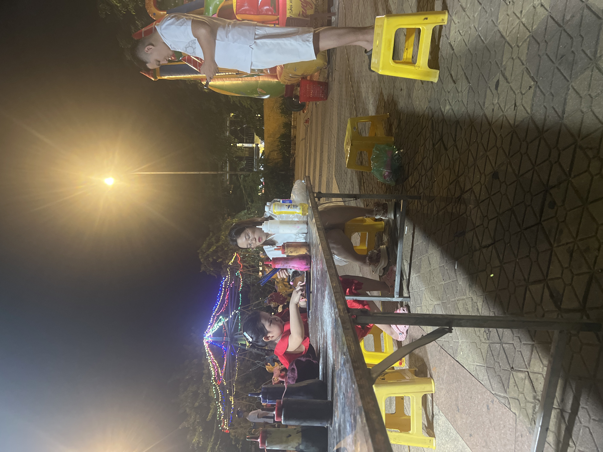At night, a girl in a red dress is painting at a table outdoors, with a woman sitting next to her. The scene appears to be in a public space in Hoi An, Vietnam, suggested by the illuminated carousel in the background and the style of street lighting. The table has bottles of paint and brushes on it. A man in a white shirt and shorts is standing to the right, looking at his phone. The ground is paved with hexagonal tiles. A large inflatable structure, possibly a bouncy castle, is visible in the far right background.