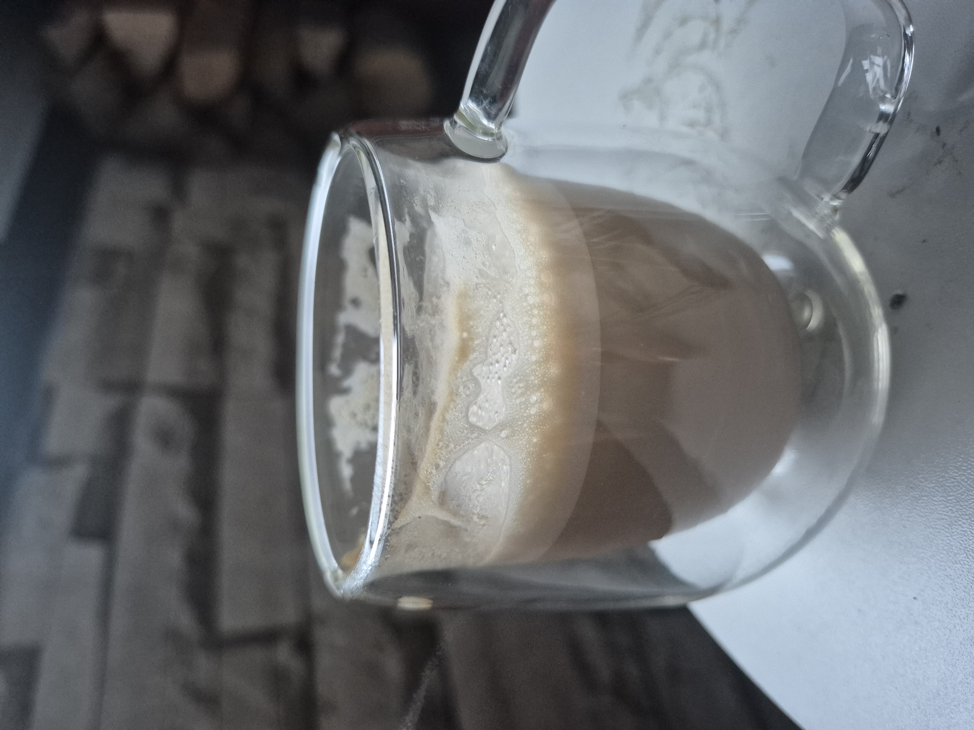 A clear, double-walled glass mug containing a light brown, possibly beige, frothy beverage, likely coffee or tea with milk, is positioned centrally in the frame. The beverage occupies approximately two-thirds of the mug's volume, topped by a layer of small, irregular bubbles adhering to the inner glass surface. A transparent handle extends from the right side of the mug. The mug rests on a light, textured, possibly white or light grey, granular surface. The background is heavily blurred, featuring a dark grey, rectangular pattern resembling brickwork on the left, and darker, indistinct shapes that may be stacked wood or stone on the right. The scene is set indoors in the city of Unknown, Unknown. No individuals are present.