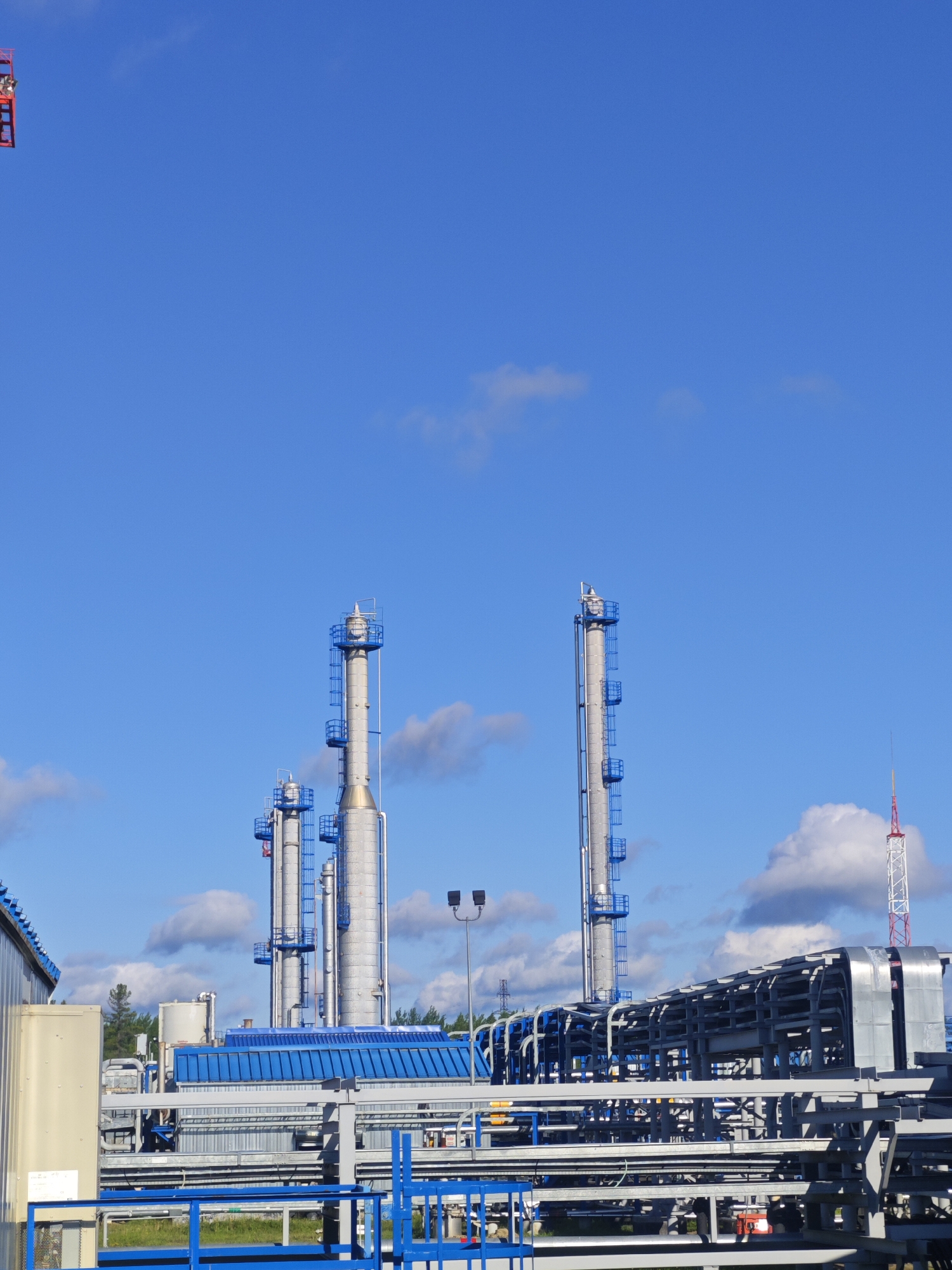 A wide, low-angle outdoor shot shows a clear blue sky with scattered white clouds above an industrial facility. In the center of the frame, two tall, cylindrical smokestacks rise against the sky, with blue railings and ladders visible on their exteriors. To the left of these stacks, a smaller, similar smokestack is partially visible. In the foreground and mid-ground, a complex network of metal pipes and structures stretches horizontally across the scene, with a blue railing in the immediate foreground. To the far left, a section of a light beige industrial building is visible, and to the far right, a red and white striped tower extends into the sky. The ground appears to be covered with grass in some areas and industrial concrete in others.