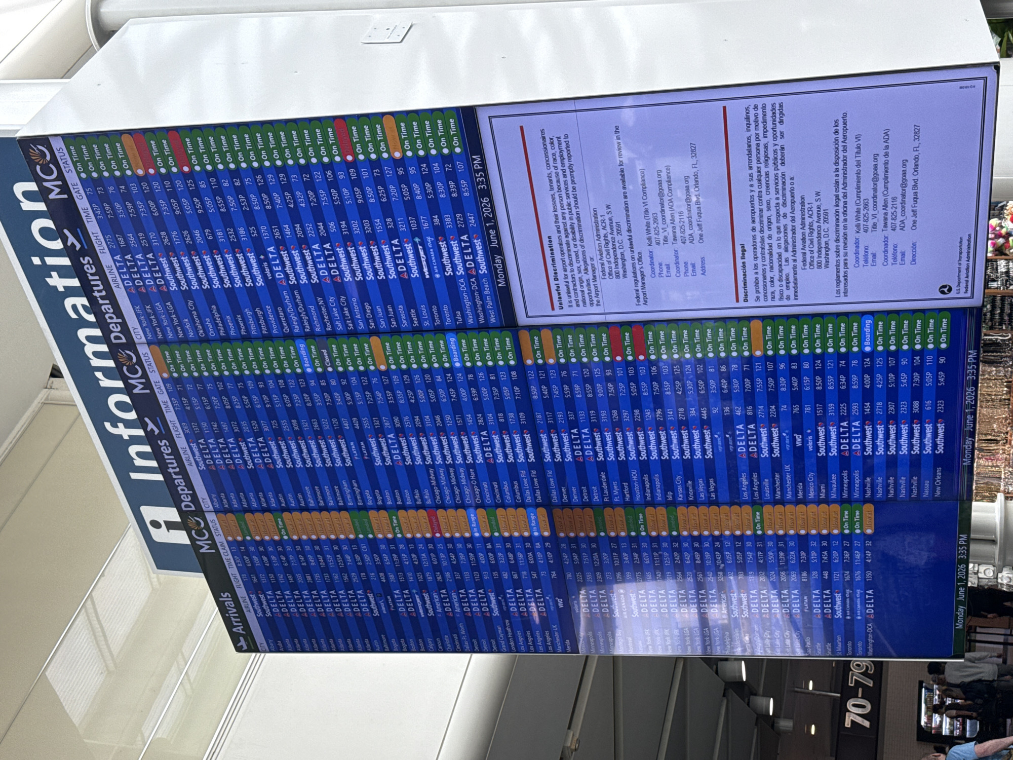 The image displays a large, vertically oriented digital flight information display board within an airport terminal. The board is divided into "MCO Information" at the top, followed by "Departures" and "Arrivals" sections. The primary activity is the presentation of real-time flight data.
The flight information is organized into columns: City, Airline, Flight number, Time, Gate, and Status. Numerous flights are listed for various airlines, including Delta, Southwest, Volaris, Air Canada, Flotam, and American, traveling to a wide range of domestic and international cities such as New York, Atlanta, Chicago, Los Angeles, Orlando, Miami, Dallas, Houston, Philadelphia, Salt Lake City, San Antonio, Seattle, Tulsa, Washington, Toronto, Montreal, Cancun, Mexico City, London, and Bogota. The majority of flights are marked "On Time," though some statuses like "Delayed," "Boarding," "Closed," "Now at Gate," and "In Range" are also visible.
A prominent feature is a legal information panel on the right side, titled "Unlawful Discrimination" in English and "Discriminación Ilegal" in Spanish. This section provides details regarding regulations against discrimination and contact information for the Federal Aviation Administration and airport ADA coordinators (Kelli White and Tawana Allen) located in Orlando, FL 32827.
The display board shows the date and time as "Monday June 1, 2026 3:35 PM" in the top and bottom right corners. The bright, indoor lighting of the airport terminal reflects slightly on the screen. In the lower right corner of the overall image, parts of what appear to be people are visible, along with gate numbers "70-79," indicating the board's placement in a passenger area.