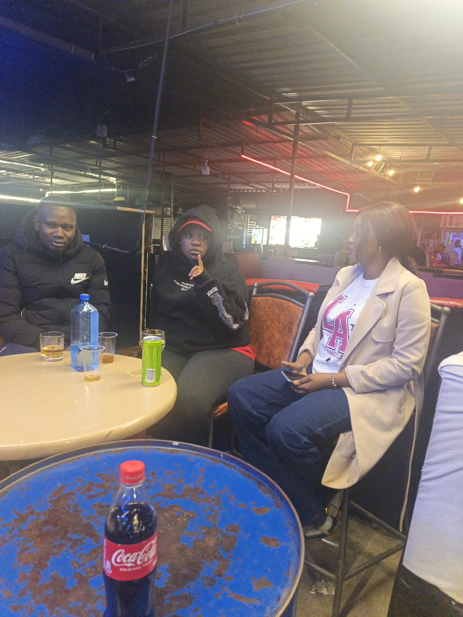 The image shows three people sitting at tables in what appears to be a bar or lounge. A man in a black Nike jacket sits to the left, with a table containing glasses and bottled drinks. A woman in a black hoodie is in the center, and a woman in a tan coat sits to the right, holding a phone. A blue table with a Coca-Cola bottle stands in the foreground. Red neon lighting is visible in the background, suggesting an urban environment. Given the description in the prompt, this location is likely in Nairobi, Kenya.