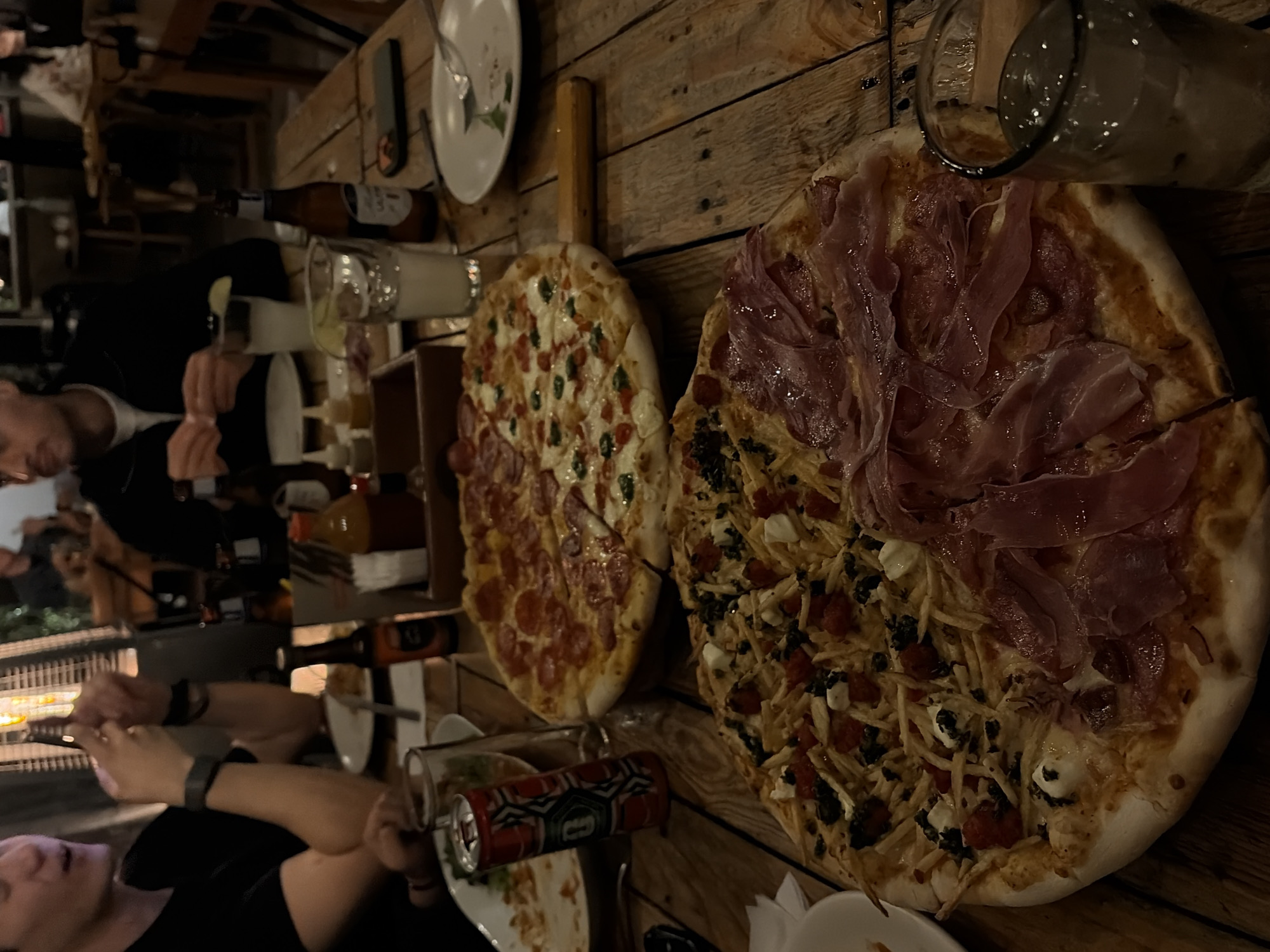 The image captures a restaurant scene with two pizzas prominently displayed on a wooden table. One pizza is topped with pepperoni and herbs, while the other is divided into halves, featuring ham on one half and a topping with potatoes, tomatoes, mozzarella, and olives on the other. A glass with a straw is positioned near the ham pizza. Dishes and cans are scattered around the pizzas, suggesting a shared meal. In the background, blurred figures indicate other diners. The overall lighting suggests an indoor, possibly dimly lit, restaurant setting. The location is Unknown, Unknown.