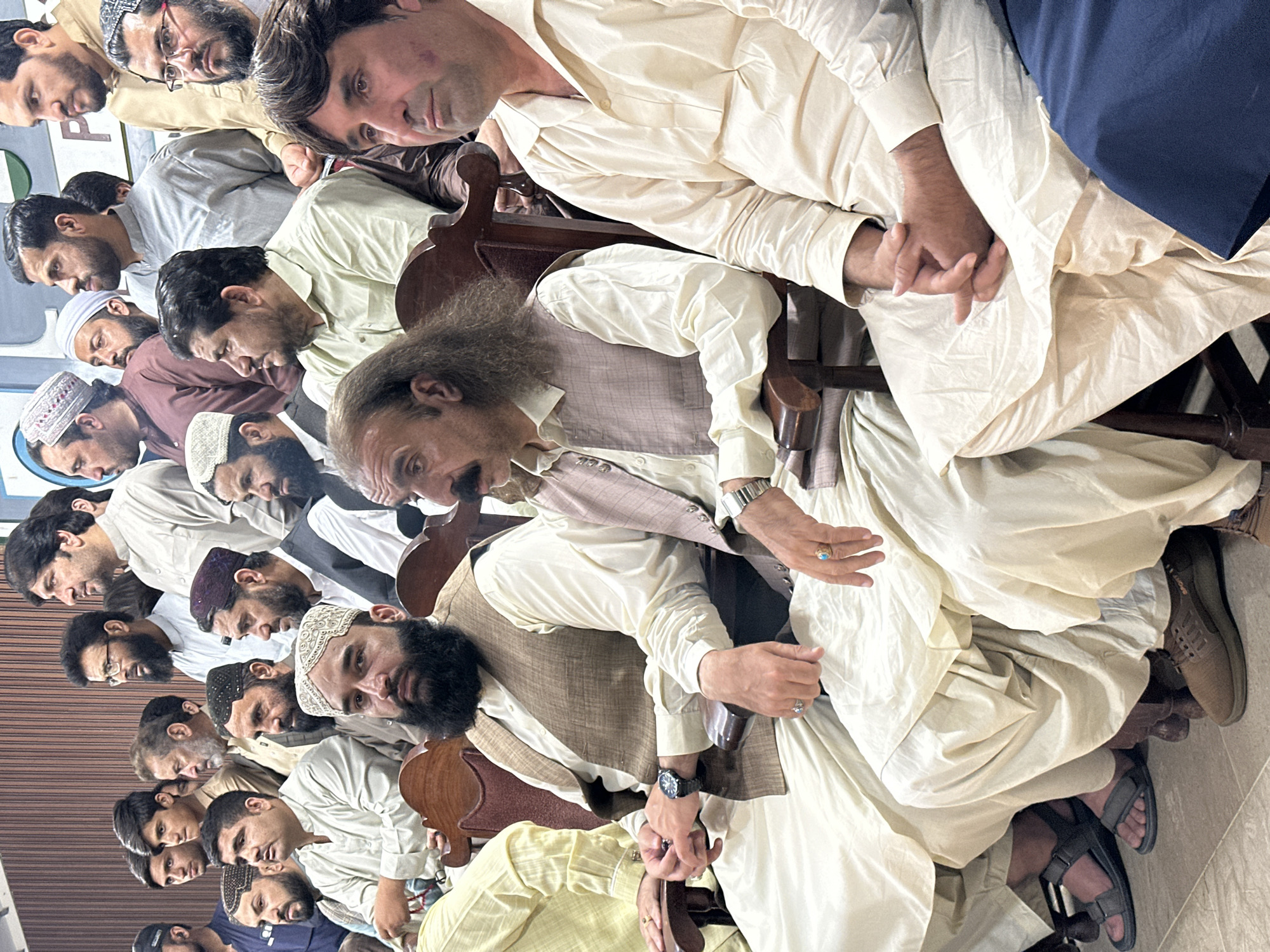 A group of men sit closely together in Quetta City Tehsil, Pakistan. They are wearing traditional clothing. Many men are wearing white prayer caps. The men are seated in chairs, and some are holding hands.