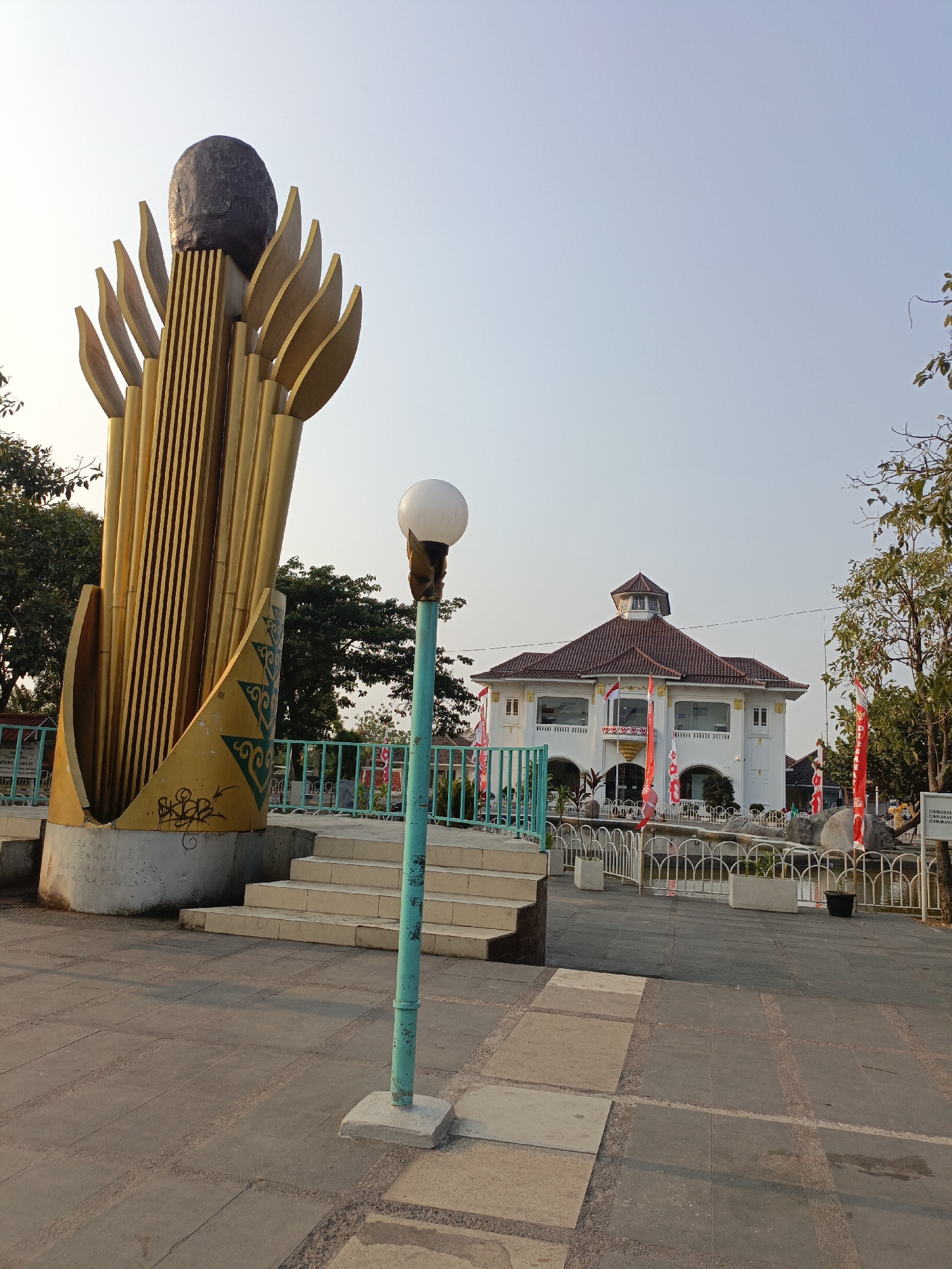 The image depicts a monument with a dark, oval-shaped top and a golden, stylized base, resembling a torch or a flame. To the right of the monument, a teal-colored lamppost with a white globe stands on a concrete base. Behind the monument and lamppost, a light-colored, two-story building with a brown tiled roof is visible. The building has a central tower with a smaller domed roof and multiple windows. Red and white flags are displayed in front of the building and along a white fence. The foreground is paved with square stone tiles, with some lighter rectangular tiles interspersed. A set of concrete stairs leads up to the monument area. Trees and foliage are present in the background. The scene appears to be an outdoor public space, likely a park or plaza. The presence of the flags suggests it might be a national holiday or a significant event. The overall setting is an urban or semi-urban environment in Tambun, Indonesia.