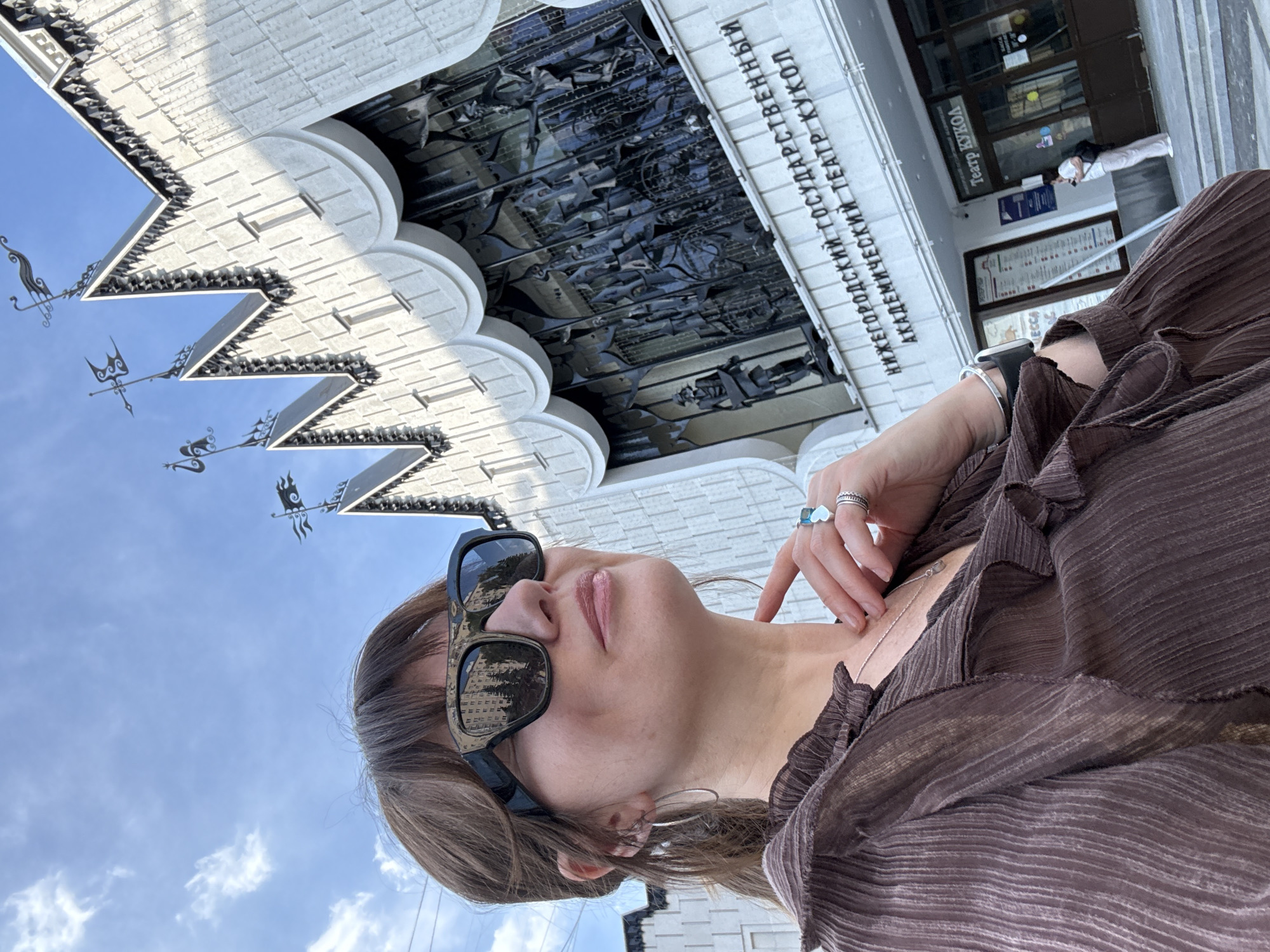 The image is a selfie taken outdoors, featuring a woman in the foreground. She is wearing sunglasses and a brown, pleated top. Her right hand is raised, with fingers touching her neck. She is adorned with jewelry, including a bracelet on her left wrist and rings on her fingers, one of which is shaped like a heart.
The background shows a large, ornate building with architectural details and relief sculptures. Text on the building reads "НИЖЕГОРОДСКИЙ ГОСУДАРСТВЕННЫЙ АКАДЕМИЧЕСКИЙ ТЕАТР КУКОЛ," which translates to "Nizhny Novgorod State Academic Puppet Theatre." There are also windows and doors visible on the building, and some posters are displayed. The sky above is blue with scattered clouds, suggesting it is daytime. The overall impression is one of an outdoor tourist photo in front of a cultural landmark.