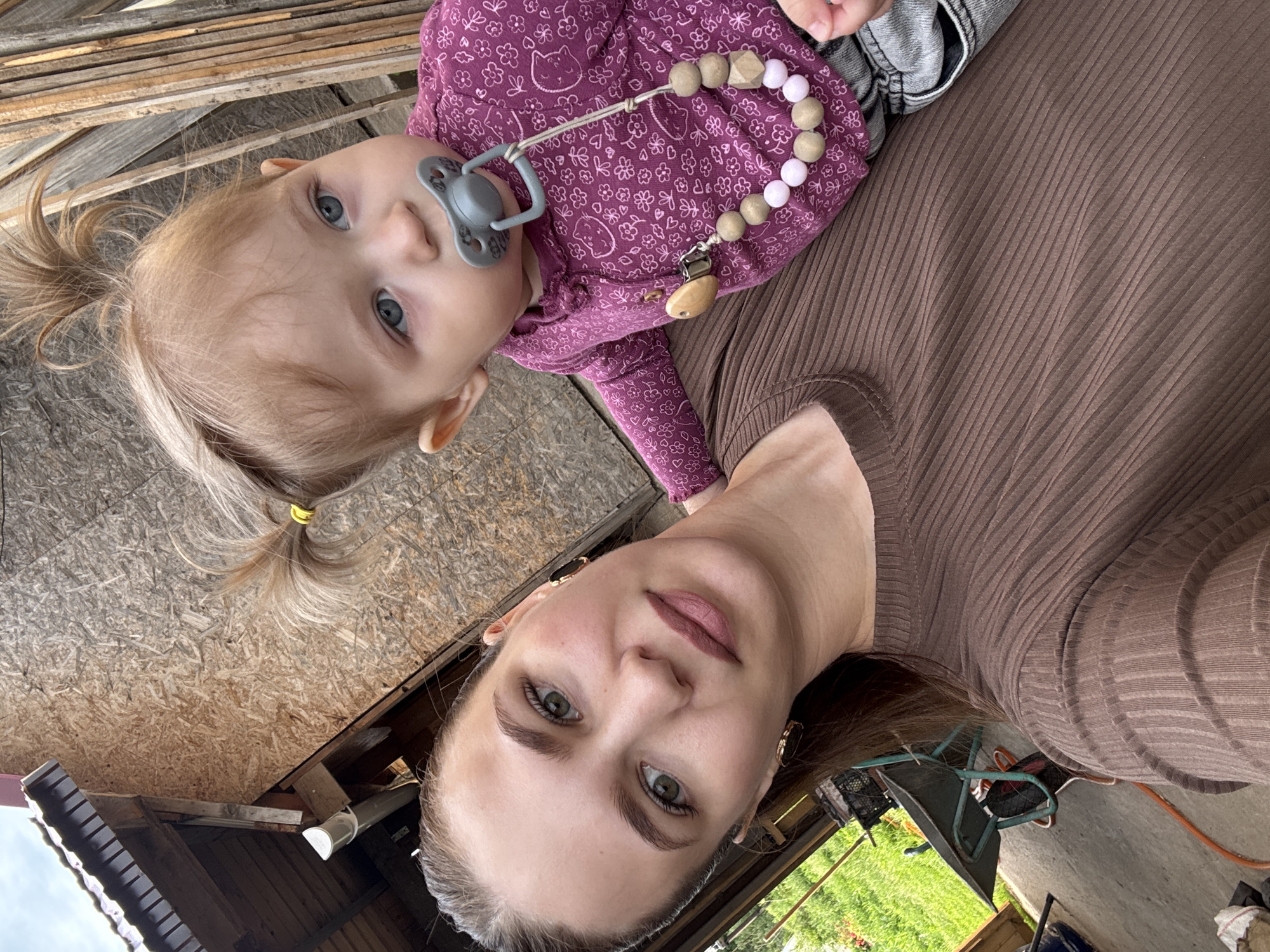 A medium shot, taken from a slightly elevated angle, captures a woman holding a young child outdoors in Nizhnyaya Gar', Russia. The image is oriented vertically with the subjects positioned diagonally.
The primary subjects are a woman and a toddler. The woman, likely the mother, occupies the lower right portion of the frame. She has light brown hair, pulled back, and green eyes, looking directly at the camera with a calm expression. She wears a brown, ribbed long-sleeved top. Her face shows light makeup, including defined eyebrows and neutral-toned lipstick.
Cradled by the woman, the toddler is positioned towards the upper left, also looking directly at the camera with wide blue eyes and a curious or neutral expression. The child has blonde hair tied into two small pigtails with yellow hair ties. They are wearing a purple long-sleeved top with a repeating pattern of white flowers and cat faces, along with grey jeans. A grey pacifier is in the child's mouth, attached to a decorative pacifier clip made of wooden and pink/grey silicone beads, ending with a wooden clip featuring an animal-like head.
The background suggests a casual, outdoor setting, possibly a backyard or a semi-finished area. Behind the child are stacked wooden planks or OSB boards. To the upper left, a patch of blue sky is visible, indicating clear weather. Parts of a wooden roof structure with a gutter can be seen in the middle left. Further into the background, hints of green grass, a metal chair, and an orange hose suggest an informal outdoor space. The natural lighting indicates it is daytime. No discernible text is visible.