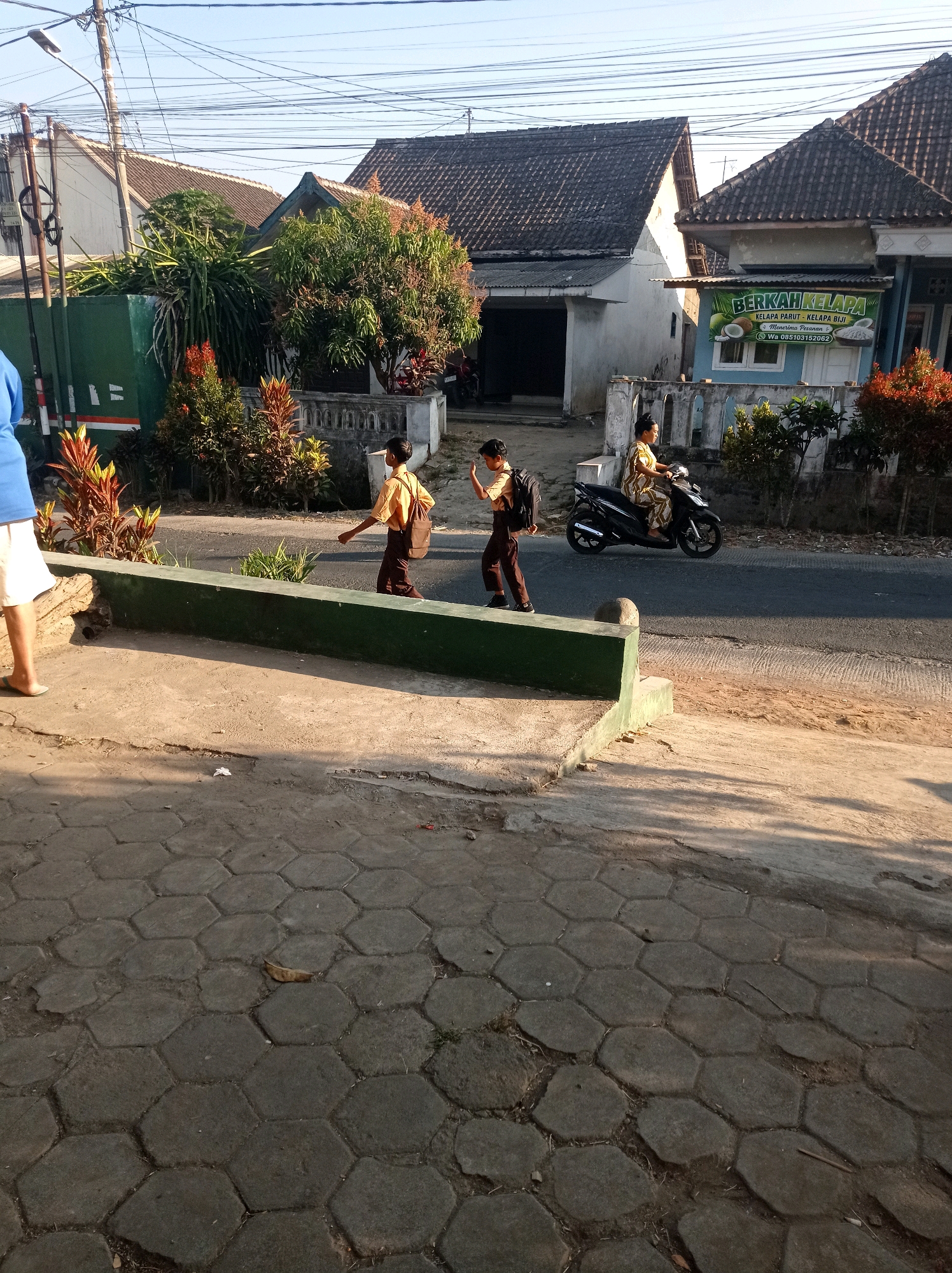 In Purwosekar, Indonesia, two young students wearing school uniforms are walking on a paved sidewalk beside a road. The student on the left, facing right, has a dark backpack slung over their shoulder. The student on the right, also facing right, is slightly ahead and also carries a backpack. A woman in a patterned dress is riding a black motorcycle in the same direction on the road. Behind the students and the road, there is a low green wall, followed by a sidewalk, and then a street with houses and trees. One house has a sign that reads "BERKAH KELAPA." The scene is bathed in sunlight, casting shadows on the ground. The sky is clear and blue.