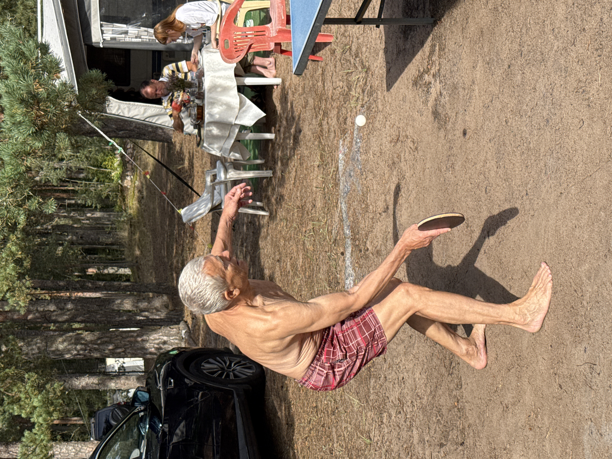A shirtless elderly man with gray hair and wearing red plaid shorts is playing ping pong outdoors. He is in mid-swing, with a ping pong paddle in his right hand. A white ping pong ball is visible on the sandy ground in front of him. In the background, a table with a white tablecloth is set for a meal, with two people sitting at it. A white tent or camper is visible behind the table. A black SUV is parked to the left of the frame. The scene appears to be a campground in a wooded area. The location is specified as Ozerki (Krasnodolinskaya), Russia.