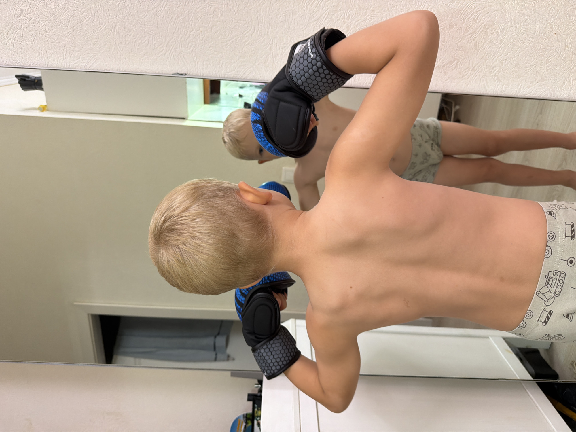 A young, shirtless boy with blond hair wearing boxing gloves is standing in front of a mirror. The boy's reflection shows him wearing patterned shorts. The scene appears to be inside a room, potentially a home. There's no clear indication of the location being in Sochi, Russia.