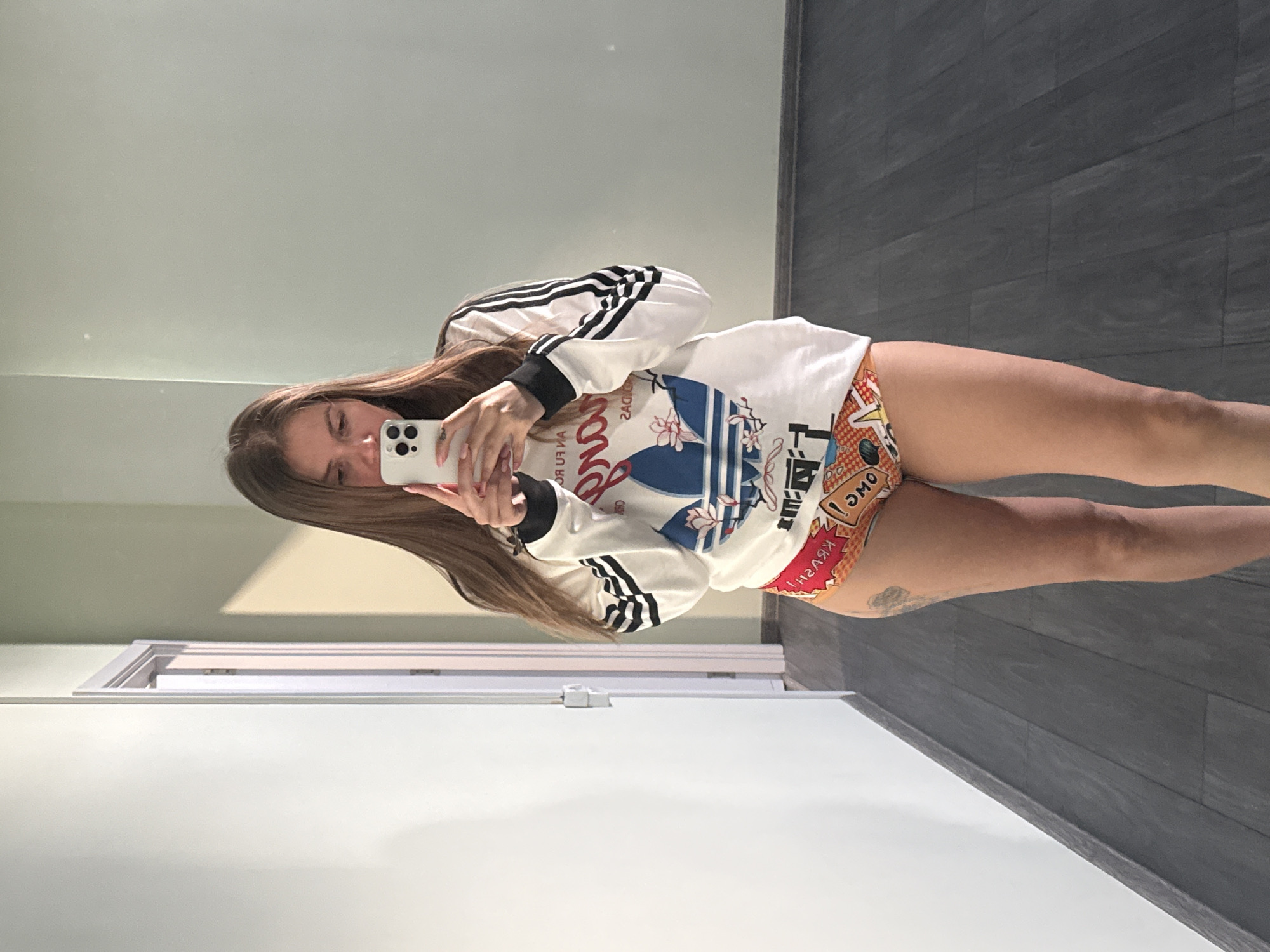 A woman is taking a selfie in a room with gray wooden floors and light green walls. She is wearing a white Adidas sweatshirt with black stripes on the sleeves and colorful printed bottoms. She holds a white smartphone in her hands, obscuring part of her face. Her long brown hair falls over her shoulders. Behind her is a white door frame and the white ceiling. It's impossible to ascertain the location in the city of Batumi, Georgia from this image.