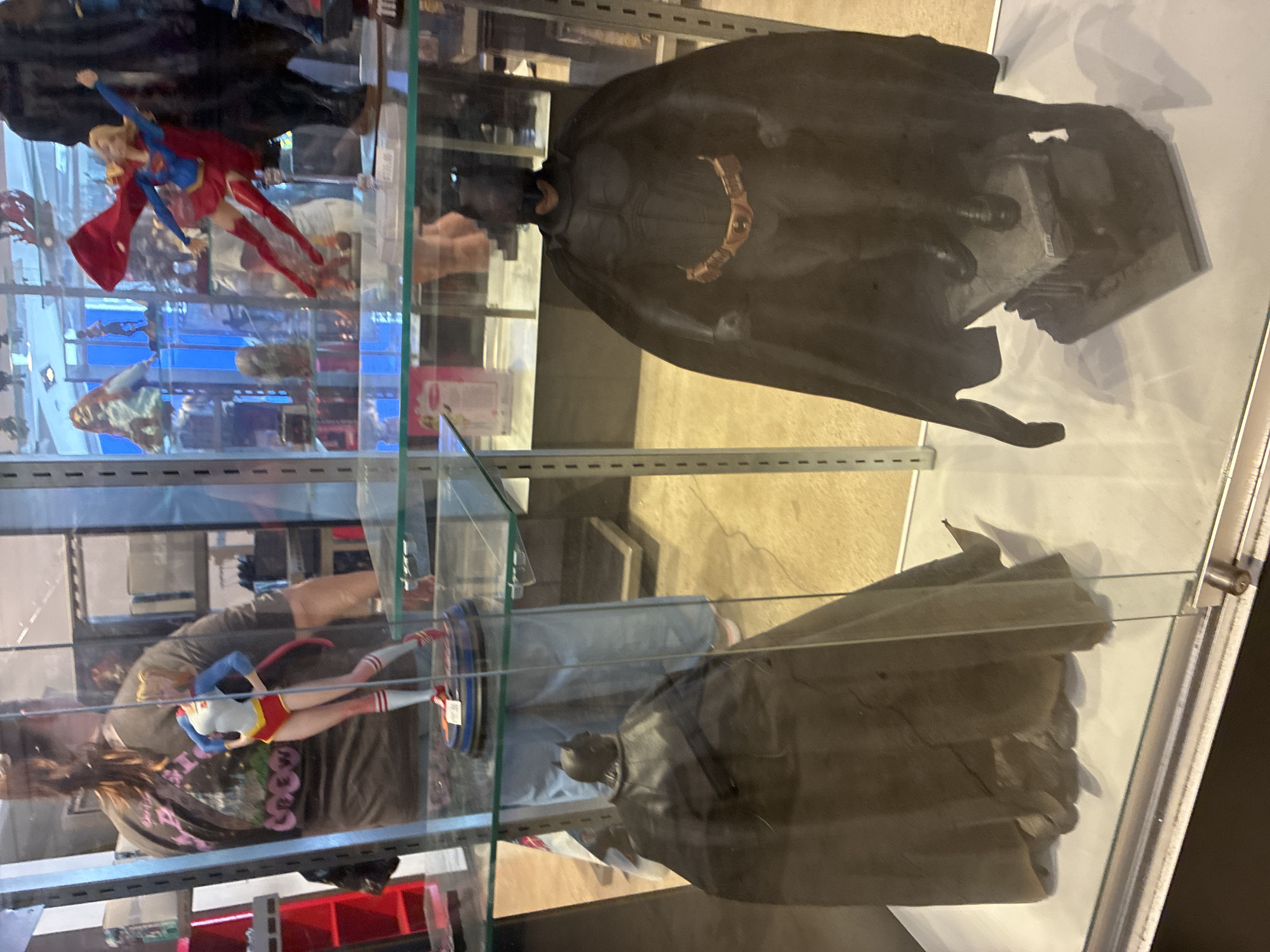 An indoor shot of a glass display case shows two Batman action figures and a Supergirl action figure. The Batman figures are on the bottom shelf, and the Supergirl figure is on the top shelf. A person wearing a dark t-shirt is visible behind the Supergirl figure. The figures are well-lit, and the background is out of focus.