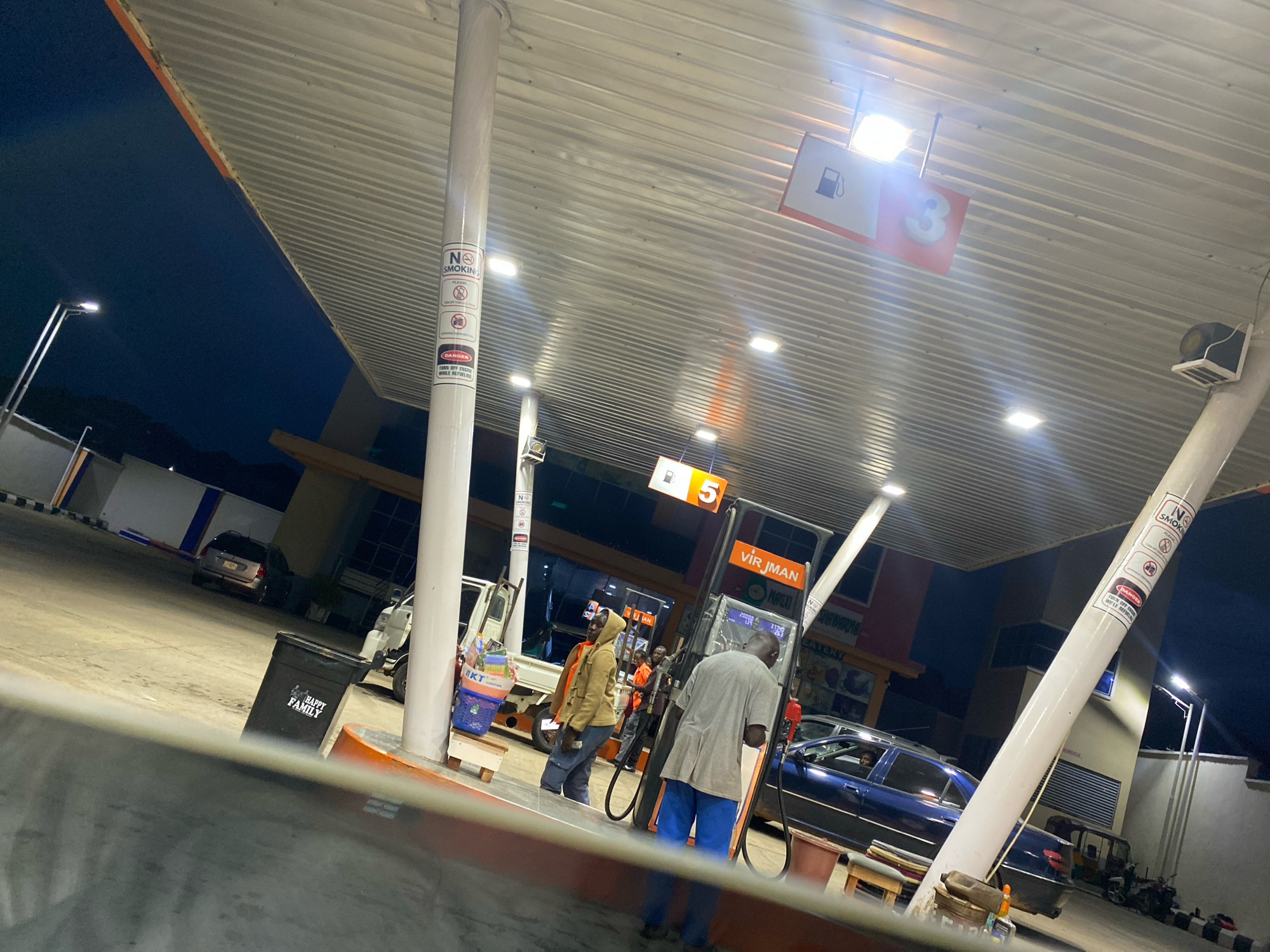 The image depicts a gas station in Jos, Nigeria, at dusk. Several individuals are present, with two men standing near a gas pump, appearing to be engaged in a transaction or conversation. One man is wearing a light brown hooded jacket and blue jeans, while the other is in a grey long-sleeved shirt and blue trousers. A dark blue car is parked to the right of the gas pump, and another vehicle, possibly a van, is visible in the background. The gas station has a covered canopy with fluorescent lights illuminating the area. Signs are posted on a pillar, including "NO SMOKING" and a "DANGER" notice. A black trash can with the text "HAPPY FAMILY" is visible on the left. The overall environment is a typical urban gas station setting.