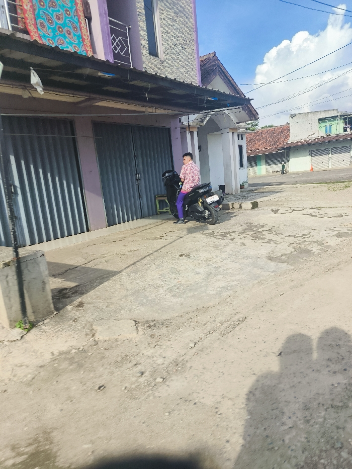 The image shows a man in a pink and white patterned shirt and purple pants sitting on a black motorcycle. The motorcycle is parked on a dirt road in front of a building with gray metal shutters. There is a blue and green fabric hanging above the shutter. The building has a stone facade and a purple awning above the shutters. There is a small building next to the larger building with a beige stucco facade and a roof with white tiles. The road is made of dirt and has a few small puddles. The location is in Sukatani, Indonesia, at the coordinates: -6.638 latitude, 107.407 longitude. The image was taken during the day with a partly cloudy sky. The scene is quiet with no other people or objects present except the motorcycle. The image is shot from a low angle and slightly skewed, giving the viewer a sense of being in the street looking up at the building. The image was taken at a slightly oblique angle. There is a small, black, metal post in the foreground.