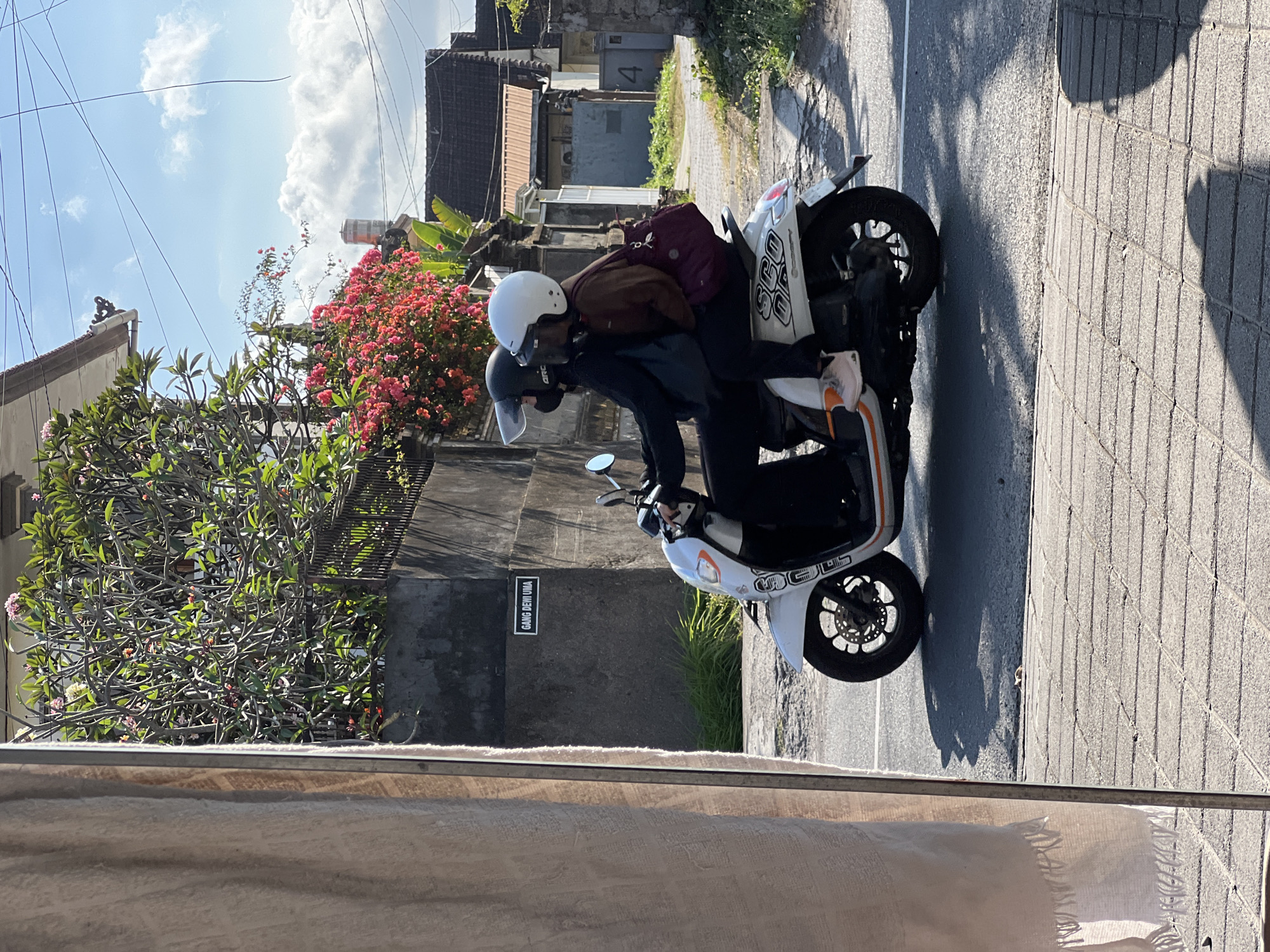 In Denpasar, Indonesia, two individuals are riding a white scooter on a paved road. The person in the front is wearing a black jacket and a black helmet, while the person in the back is wearing a brown long-sleeved shirt and a white helmet. They are both wearing masks. The scooter has the word "SCOOPY" written on its side. Behind them, a concrete wall displays a sign that reads "GANG DEWI UMA". Lush green foliage and vibrant pink flowers are visible on top of the wall. The sky is a clear blue with scattered clouds. Buildings with tiled roofs are present in the background. A large, textured beige fabric hangs on the left side of the frame, partially obscuring the view.