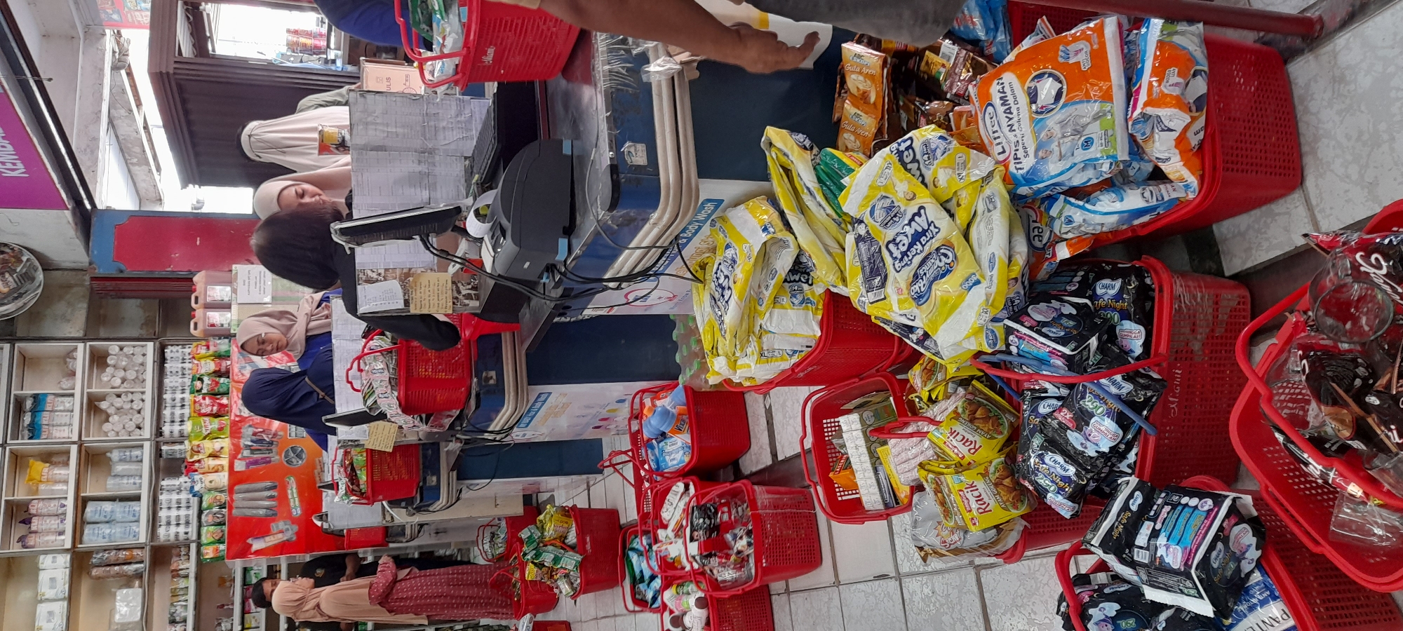 The image shows a retail store in Pemalang, Indonesia, likely a minimarket or a small grocery store. The scene is dominated by stacks of red plastic baskets overflowing with various consumer goods, primarily packaged food items and household products. The store appears to be busy, with several customers, mostly women wearing hijabs, interacting with staff at a checkout counter. The checkout counter itself is equipped with a computer monitor, a printer, and a keyboard. Shelves stocked with more products are visible in the background. The overall impression is that of a bustling commercial environment.