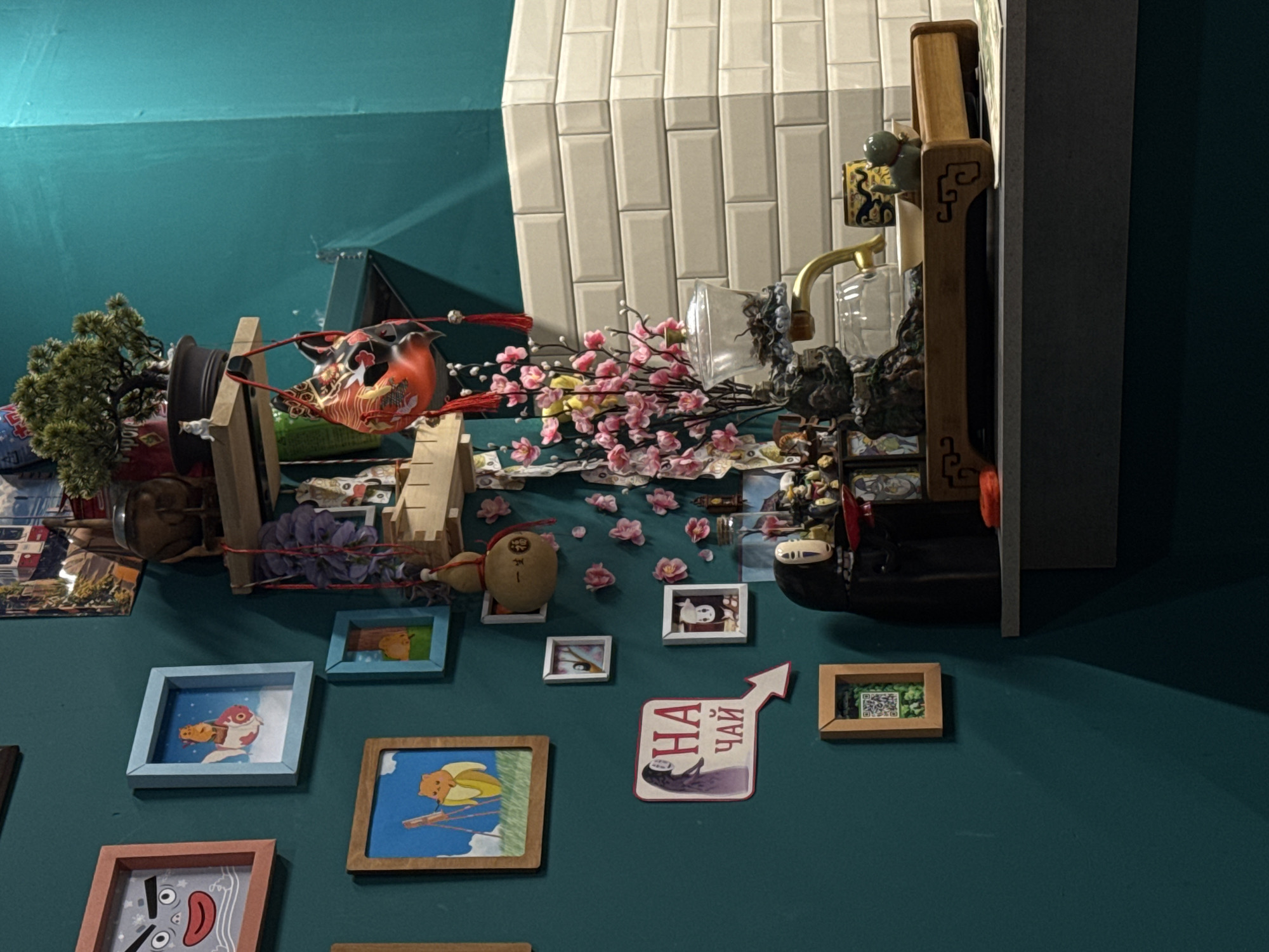This image shows a cluttered wall with various framed pictures and decorative objects in what appears to be a building in Izhevsk, Russia. A mask resembling a fox, accompanied by artificial flowers, is placed atop wooden structures. Beneath these, several small framed pictures hang on the teal-colored wall. Other items include a kettle, a QR code print, and a sticker in Russian text. The scene is bathed in artificial light.