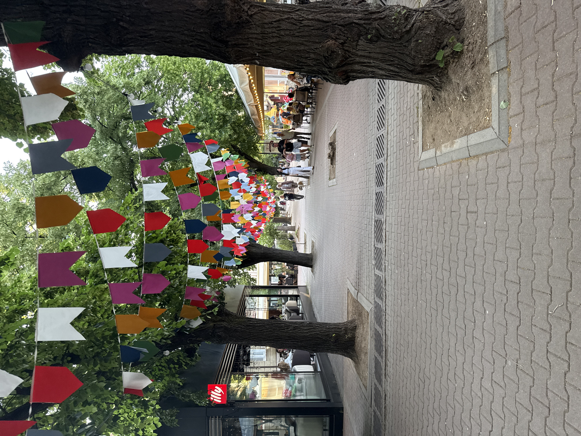 The image depicts a pedestrian street in Kyustendil, Bulgaria. The street is paved with grey interlocking paver bricks, featuring integrated drainage grates. Mature trees with dark, textured trunks line both sides of the thoroughfare, their dense green foliage forming an overhead canopy. Strung between the trees are numerous lines of pennant-shaped bunting in multiple colors, including red, white, pink, yellow, blue, green, grey, and orange, interspersed with small spherical lights. On the left, a building with large glass windows houses a cafe, marked by a red 'illy' logo; several individuals are visible seated inside. On the right, further down the street, an outdoor cafe with an awning has patrons seated at tables. In the mid-ground, a group of approximately five individuals is walking towards the background, including a person in white attire walking a small light-colored dog on a leash. Additional pedestrians are visible in the distance.