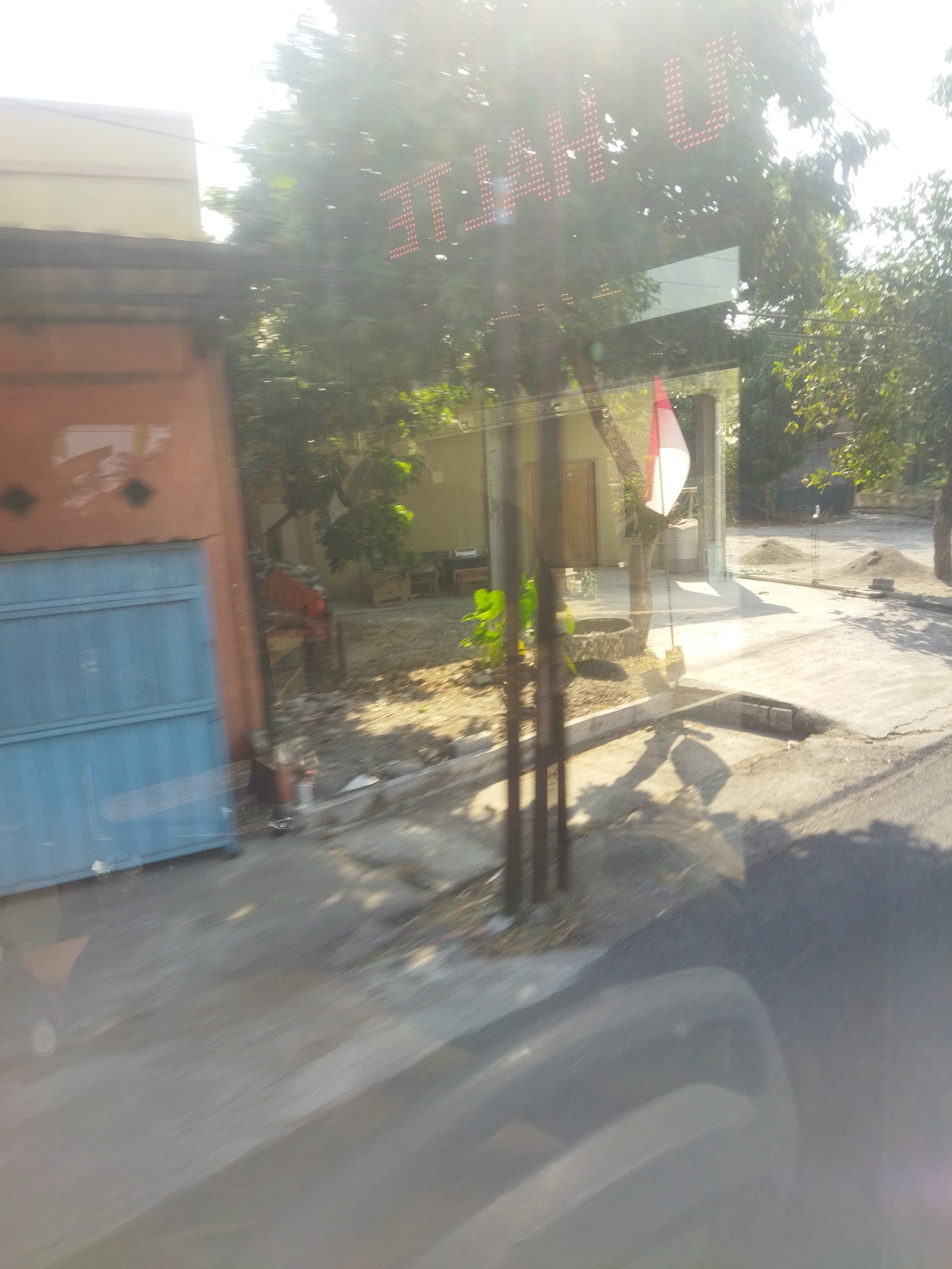 The image captures a scene from a moving vehicle, evident from the motion blur, likely in the city of Nambangan, Indonesia. In the foreground, the blurred reflection of a vehicle's interior, possibly a dashboard or seat, is visible. The middle ground reveals an outdoor scene with a coral-colored building on the left, featuring a light blue metal door. Adjacent to it, a dirt path leads to a small building with a light-colored facade and a wooden door. A flagpole stands prominently, displaying a red and white Indonesian flag. The background is dominated by lush green trees and vegetation, suggesting a partly rural or suburban environment. Sunlight is strong, causing overexposure and glare in the upper portion of the image. The overall impression is one of a typical roadside view in an Indonesian town.