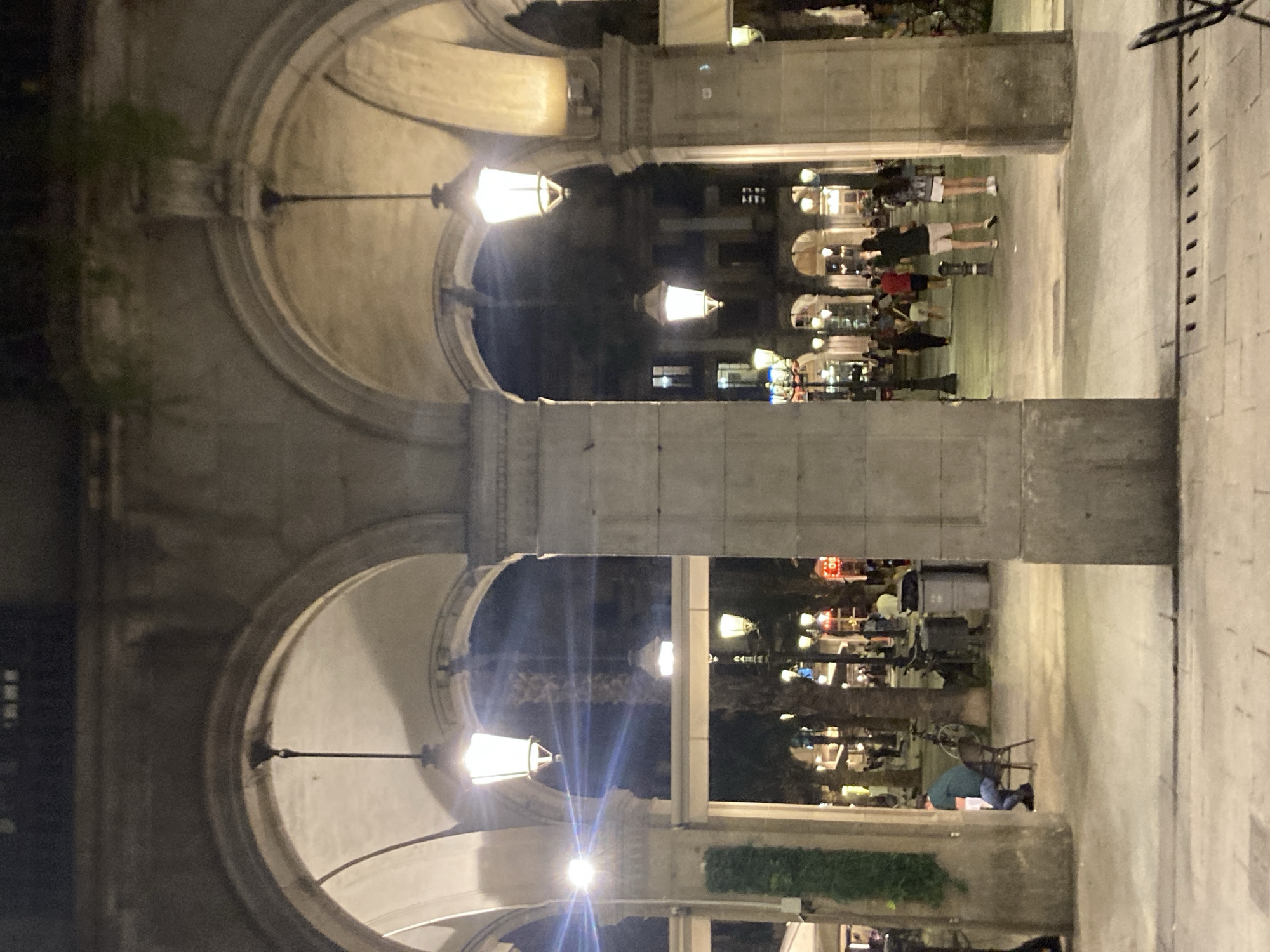 The image shows a nighttime scene under large stone arches in Barcelona, Spain, likely in a plaza or pedestrian area. Several classic street lamps are illuminated, casting a warm glow on the stone architecture and the walkway. In the foreground, two large stone pillars support the arches, framing the view.
Through the arches, a number of people are visible walking and standing. Some appear to be patrons of outdoor cafes or restaurants, as suggested by tables and chairs. The background shows buildings with illuminated windows, indicating an urban setting. Palm trees are also visible, a common feature of Barcelona's streetscape.
On the left side of the image, a person sits alone at a table, facing away from the camera, while a bicycle is parked nearby. Further into the plaza, groups of people are strolling. On the right, two individuals are walking towards the camera, dressed in casual summer attire. The overall atmosphere is one of a lively evening in a European city.