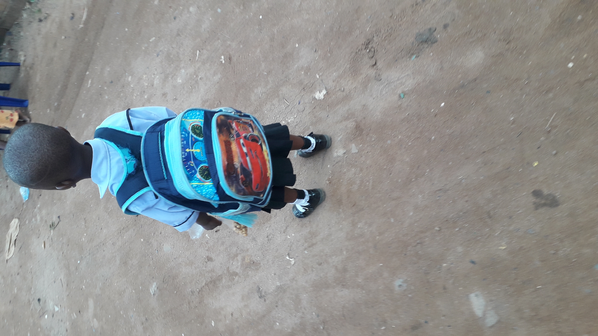 A young child stands on a rough, dirt-colored surface, presumably a street or walkway in Nkpor, Nigeria. The child wears a school uniform consisting of a white collared shirt, a dark skirt or shorts, white socks, and black shoes. A backpack with a "Cars" character design is strapped to the child's back. The child's head is shaved close to the scalp. The child holds something in their hands, but the object is not clearly visible. The background is blurred, focusing on the child.
