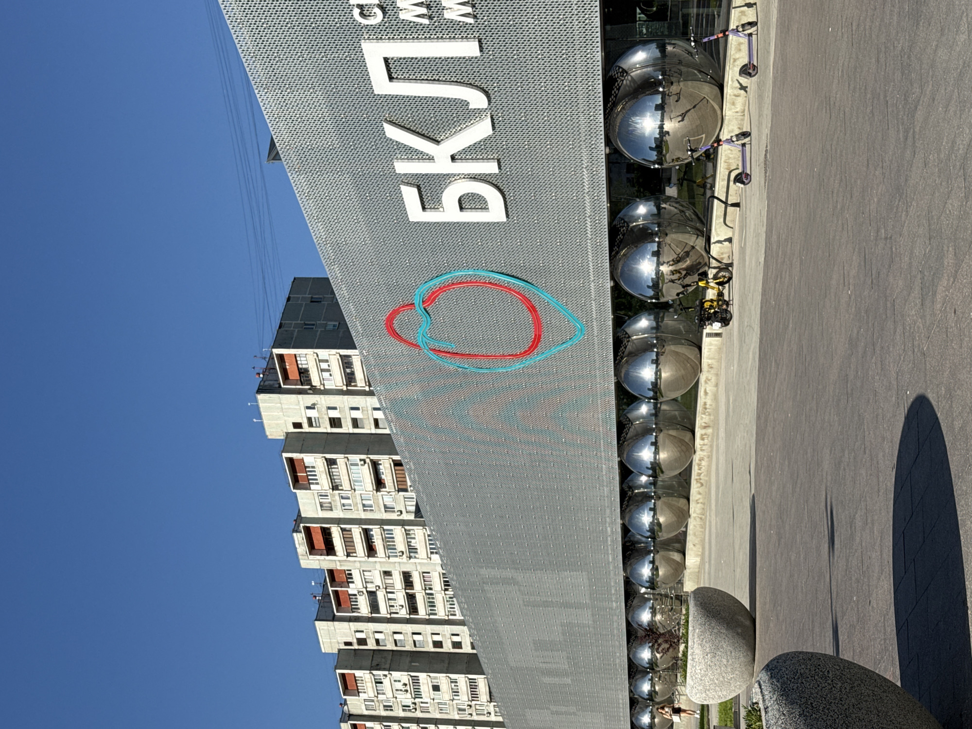 This image captures the exterior of a modern building, identifiable as a Moscow Metro station on the Big Circle Line (Большая кольцевая линия, БКЛ) due to the prominent Cyrillic letters "БКЛ" and the intertwined red and blue/teal heart-shaped logo on its perforated gray facade. Below "БКЛ," the partial letters "СМ" are visible, likely indicating "Станция Метро" (Metro Station).
The building features a long, modern design with a textured, perforated gray panel facade. Along its base, a continuous row of large, highly reflective chrome spheres is present, mirroring distorted views of the surroundings, including the blue sky, the residential buildings in the background, and the ground.
In the background, a series of multi-story residential buildings with a utilitarian architectural style, characteristic of Soviet-era panel blocks, are visible under a clear blue sky. These buildings are light-colored with some red or orange accents on balconies or window frames. Overhead utility lines stretch across the sky in the mid-ground.
The foreground consists of a paved plaza with dark gray square tiles. On the left, two large, round, light gray concrete planters containing green foliage are positioned. A person, appearing to be a woman in dark shorts and a light top, is walking away in the distance near the planters. On the right, a yellow moped or scooter with a rear cargo box is parked alongside two purple electric scooters near a bike rack, all casting distinct shadows from the bright sunlight. The scene is illuminated by strong, direct sunlight, creating sharp shadows, indicating a clear, sunny day.