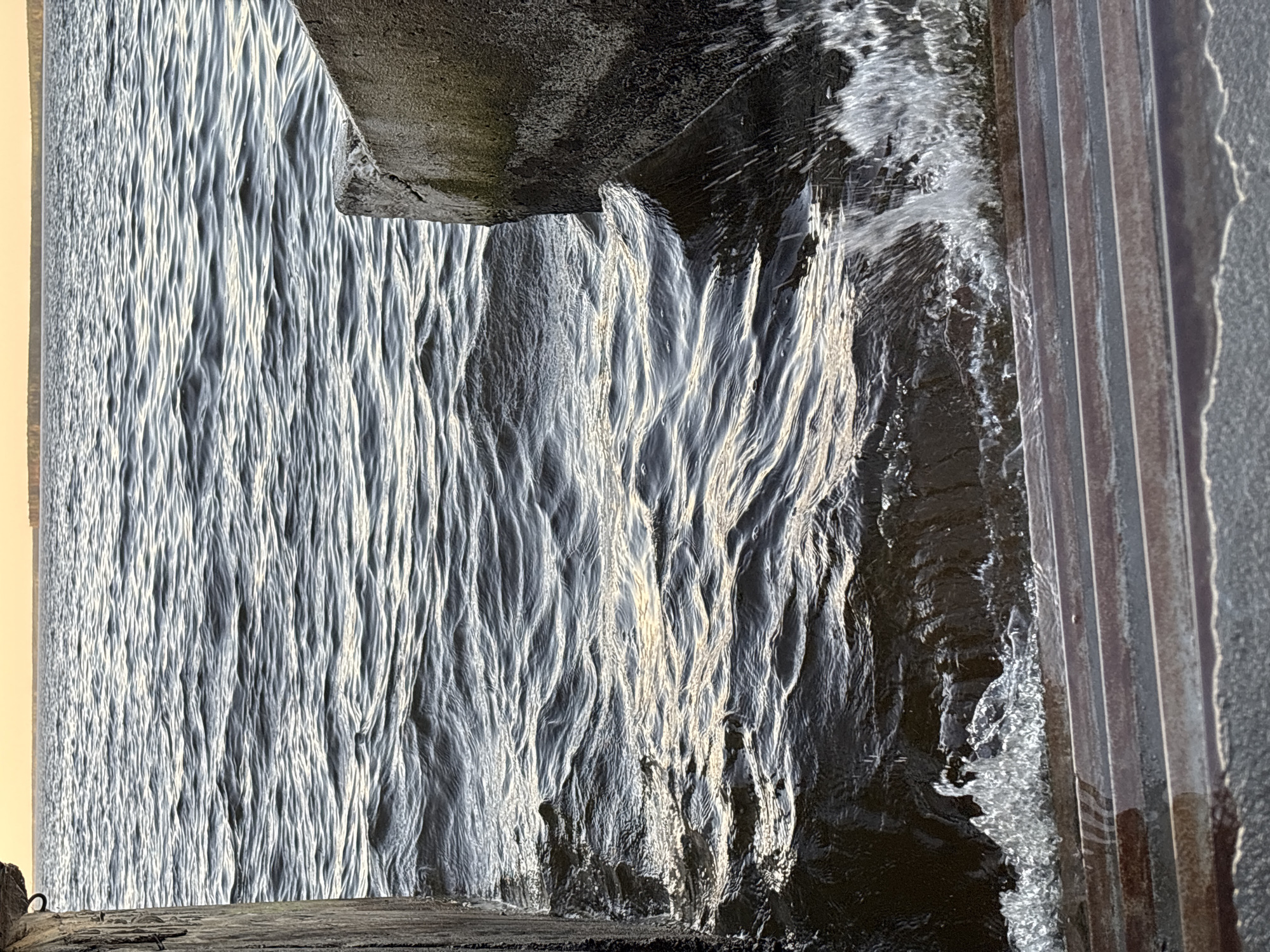 The image shows water flowing down a structure, likely a dam or spillway. The water is textured with vertical streaks and highlights, suggesting movement and sunlight. The top and bottom of the frame include portions of the concrete structure. There are metal rods or pilings on the right side of the image adjacent to the water, and possible wooden structure elements on the left side of the image.
There is no indication of the location being in the city of Chaikovsky, Russia or evidence of any people, specific subjects, objects, actions, interactions in this image.