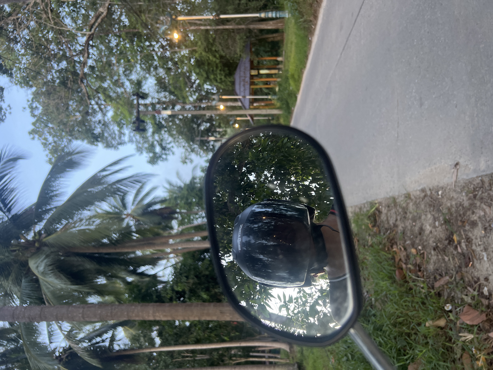 In a rearview mirror of a motorcycle, a person wearing a black helmet is visible, facing away from the camera. The mirror reflects a road flanked by lush green vegetation, including tall palm trees. In the distance, a traditional Thai-style pavilion with a dark roof is situated amongst the trees, and several streetlights line the side of the road. The scene appears to be set in a tropical location, likely Koh Phangan, Thailand, given the vegetation and architecture. The lighting suggests it might be early morning or late afternoon.
