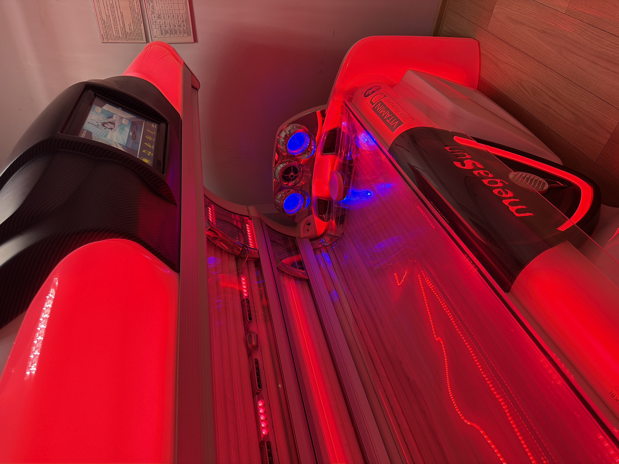 A tanning bed with red and blue lights illuminates the interior of a room. The tanning bed has a sleek, modern design with black carbon fiber accents. The lid of the tanning bed is open, revealing the padded surface where a person would lie. There are several control panels and screens visible, as well as what appears to be a cooling system with blue lights. The overall impression is one of advanced technology designed for personal care. The location is not specified in the image.