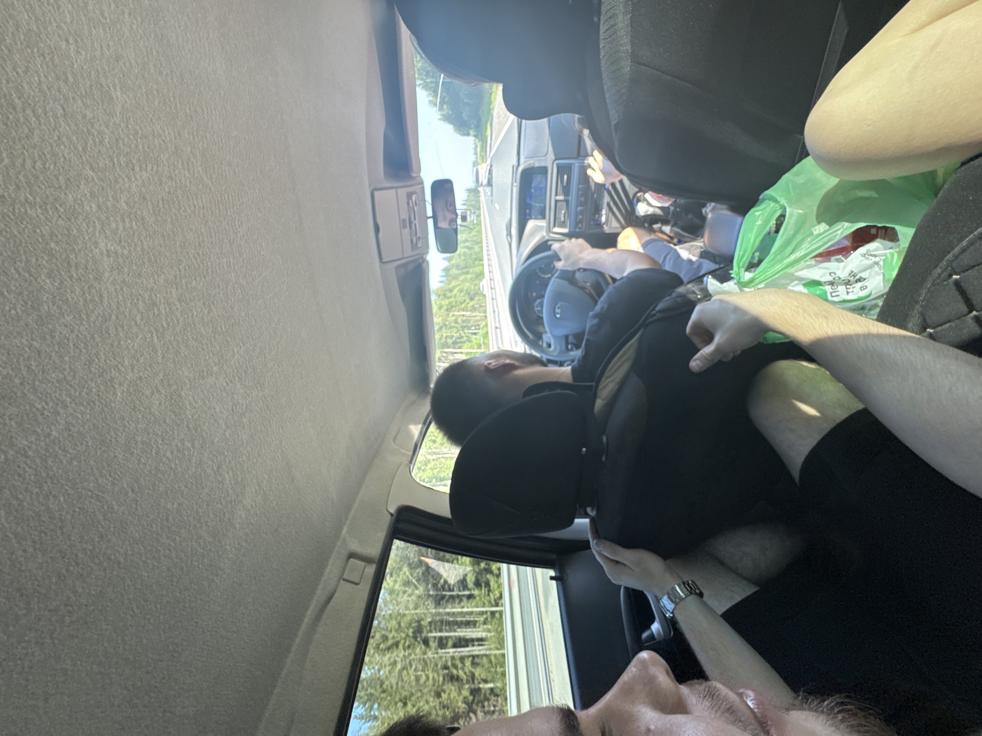 The image shows the interior of a car driving on a road, with a view of a forest on the side. Three individuals are visible: a driver at the front, and two passengers in the back. The driver is a man wearing a dark shirt, operating the steering wheel. To his right, a passenger, also a man, is partially visible from the back of his head. In the foreground, another passenger, a man, is seen from the side of his face, looking towards the camera, with his hand resting on the car's interior. A green plastic bag filled with items is located on the floor of the back seat. The car is traveling on a paved road, with other vehicles visible in the distance. The overall scene suggests a journey through a rural or semi-rural area, possibly on a sunny day given the lighting. There is no specific indication of the city of Ivanovka, Russia.