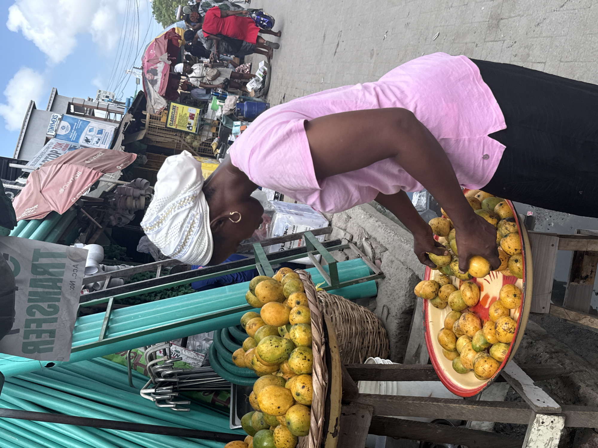 An eye-level shot reveals a woman selling mangoes at a roadside stand in Lagos, Nigeria. She is light-skinned with dark hair and is wearing a white headscarf, a pink polo shirt, and black pants. She is bending over her merchandise, picking mangoes from a yellow-and-red tray. Two baskets are crammed with mangoes of varying ripeness; some are a vibrant yellow, while others are still partially green. The stand itself is a makeshift arrangement of wood. To the left of the woman is a stack of green PVC pipes and metal hangers.
In the background, the bustling street of Lagos can be seen. Numerous people are walking by, and vendors have set up shop under umbrellas and makeshift shelters. Signs for various businesses, including "TONYMEKS" and "CLASSICLINK COMMUNICATION," are visible.