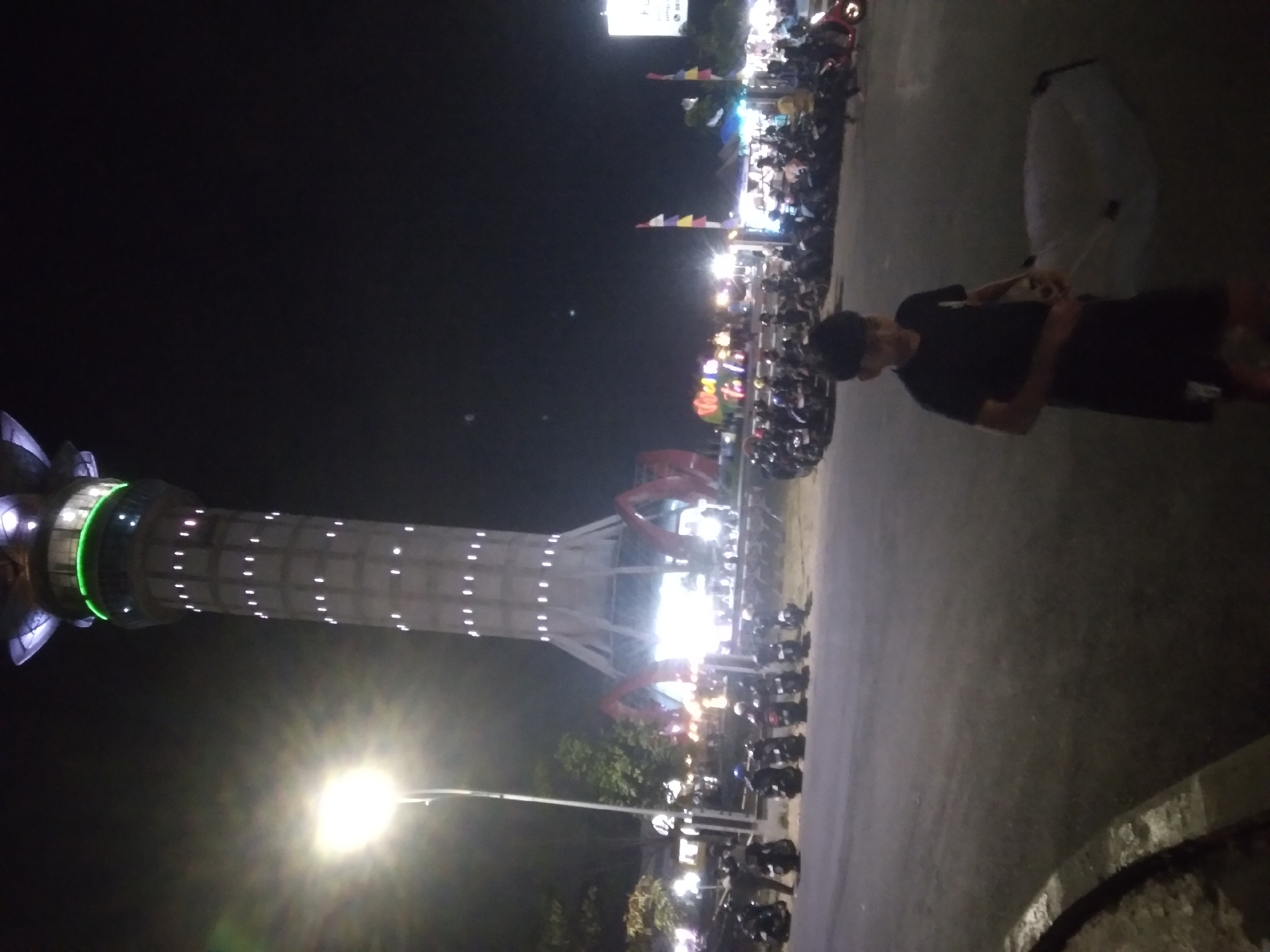 The image depicts a nighttime scene in Purwokerto, Indonesia, likely near the Tugu Pahlawan (Heroes Monument) or a similar landmark, given the prominent tower in the background. A tall, cylindrical tower with numerous illuminated windows stands illuminated against the dark sky. The tower's base is supported by angled structures, and decorative lights, possibly green, encircle a section near its top.
In the foreground, a young boy is seen walking. He is wearing a black t-shirt and black shorts and appears to be holding a white, circular object, possibly a kite or a decorative item. Behind him, a paved area shows parked motorcycles, indicating a public gathering or event. Further in the background, there are signs of commercial activity, with illuminated stalls or shops and more people. Flags with colored horizontal stripes are visible on poles, adding to the festive atmosphere. The overall impression is of a lively urban environment at night, with the tower serving as a central point of interest.