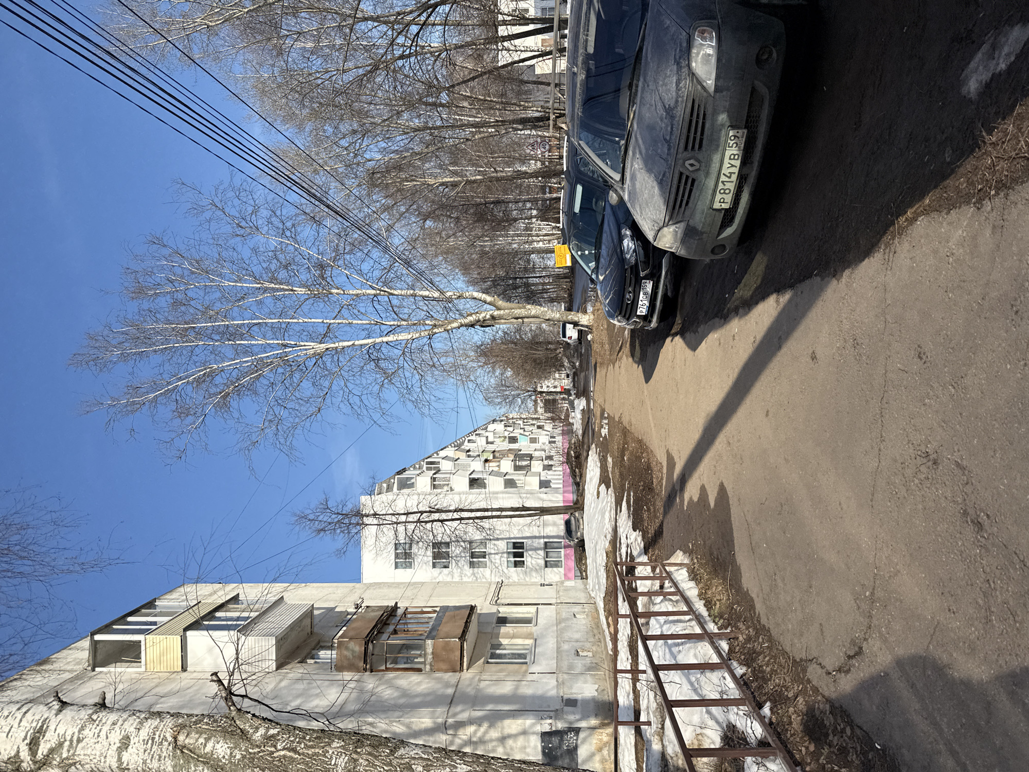 The image captures a street scene in Chaikovsky, Russia. The view is along a sidewalk adjacent to a road where two cars are parked. The first car, a dark-colored sedan, is only partially visible and obscured by the second, larger, gray/dark blue Renault Logan. The Renault's license plate reads "P 814 YB 59". Alongside the road are leafless trees lining the street.
On the other side of the sidewalk are gray buildings. The first building is a low apartment complex with protruding window boxes and a small metal fence with remnants of snow visible on the ground. The second building is taller and white, with many windows, some of which appear to be attic windows. Overhead, a network of electrical wires is visible against a clear, blue sky.