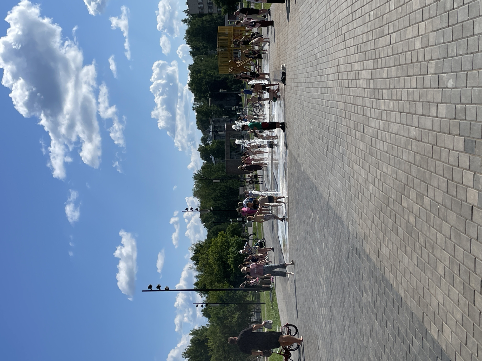 A full, outdoor, eye-level shot shows a crowd of people, many of them children, enjoying a splash pad on a bright, sunny day in Kirovo-Chepetsk, Russia. The splash pad is in the center of the frame, with water jets spraying upwards. Children are running through the water, laughing and playing. Adults are standing around, some watching the children and others interacting with them. The ground around the splash pad is paved with gray interlocking bricks, and a few bicycles are parked nearby. In the background, there are trees, buildings, and a playground structure. The sky is blue with fluffy white clouds. The overall scene is one of joy and recreation.