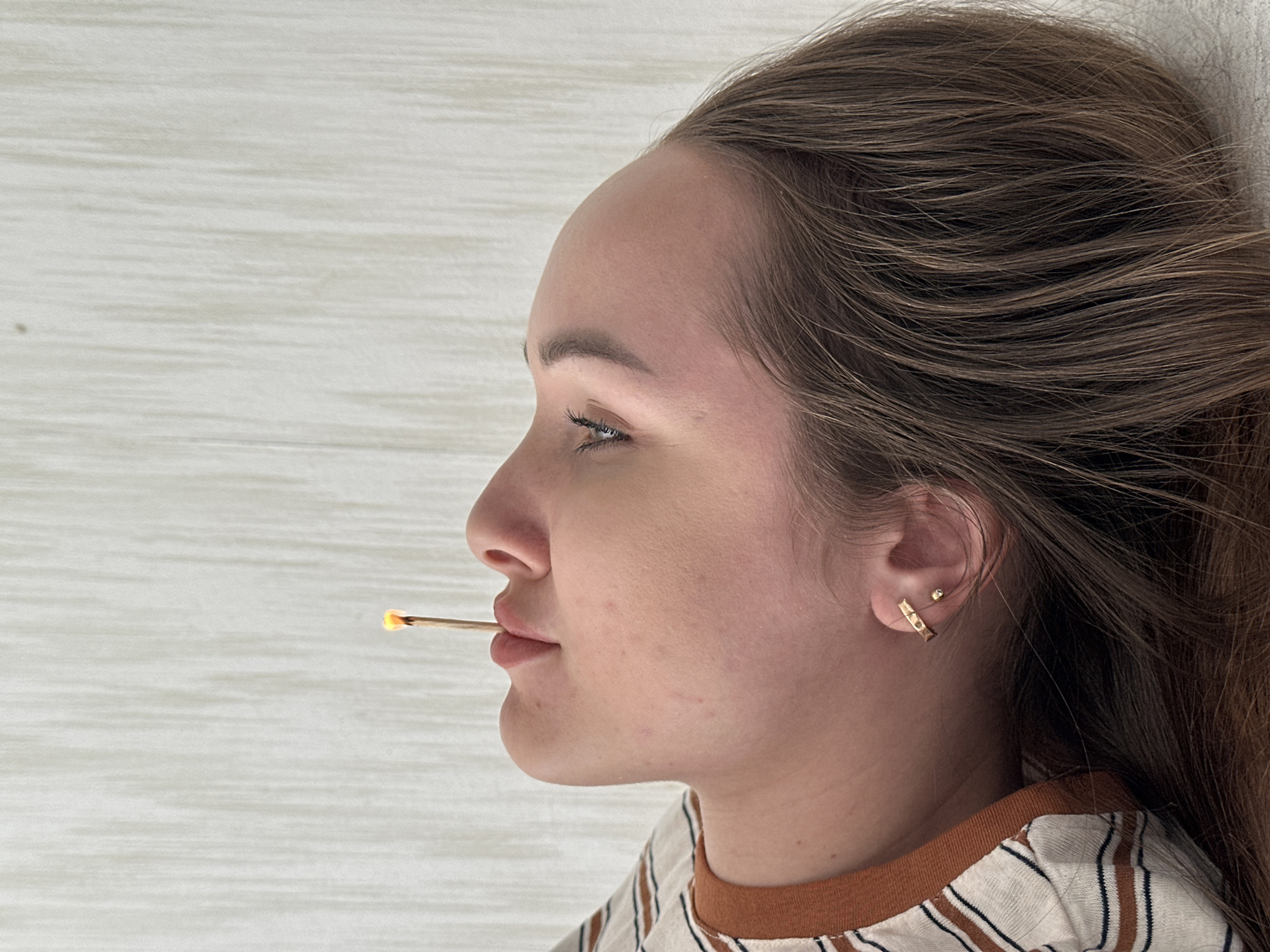 A profile shot depicts a young woman holding a lit match between her lips. She is lying on a pale, horizontally-grained surface, possibly a wooden floor or table. Her hair is brown with lighter highlights, and she has fair skin with minimal makeup. She wears a striped shirt with a brown collar and small gold earrings. The location cannot be determined from the image and there is no indication that it is from Chaikovsky, Russia.