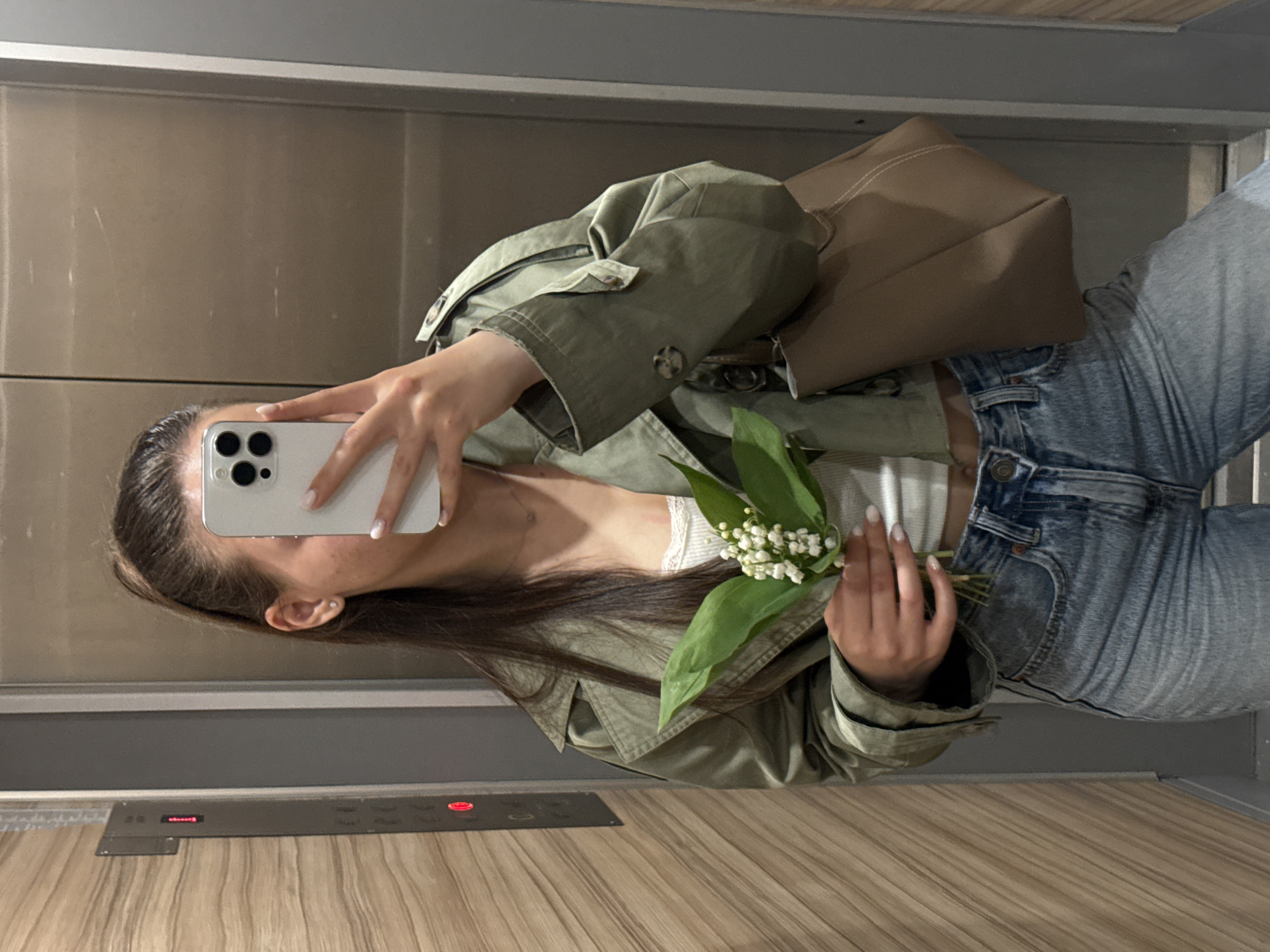 A medium shot shows a woman with long dark hair in an elevator, taking a photo of herself with a white smartphone. She is wearing a light green jacket, jeans, and a beige handbag hangs off her shoulder. She is holding a bouquet of lily of the valley flowers. The elevator's control panel is visible near the bottom of the frame. The location is not identifiable as Sofia, Bulgaria.