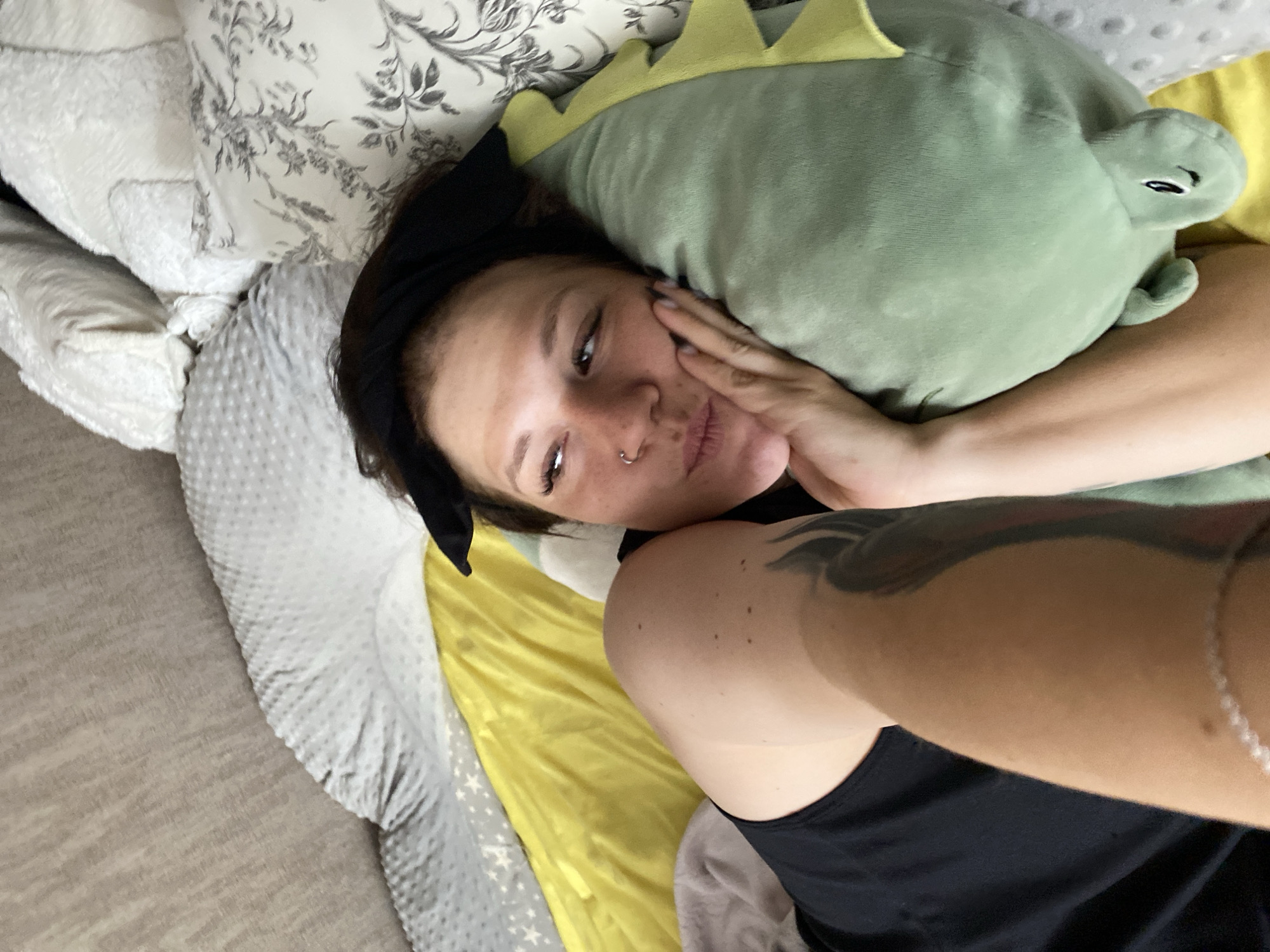 A woman with a black headband is lying on a bed with a dinosaur-shaped pillow. Her right arm is resting on the pillow, and her head is propped up by her hand. She is wearing a black tank top. The bed has several pillows, some with a gray, dotted pattern and others with a floral pattern. A yellow sheet is visible behind her. The scene appears to be indoors, likely in a bedroom. There is no information about the location in Chaikovsky, Russia.