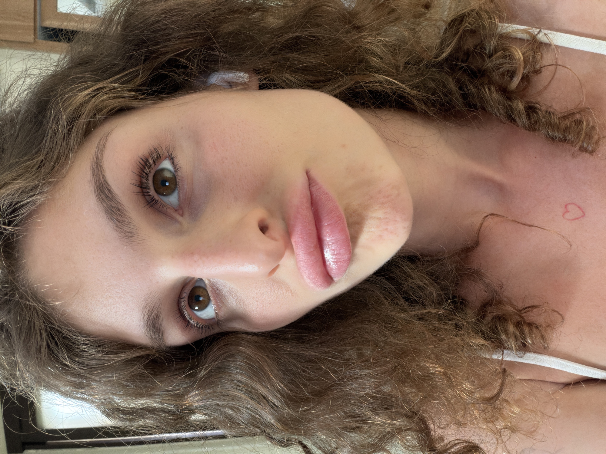 A close-up portrait of a young woman with curly brown hair. She has fair skin, brown eyes, and full lips. Her expression is neutral. She is wearing a white top. There is a small red heart tattoo on her upper chest. The image is shot indoors. There is no information in the picture to confirm that this image has any relation to North Kuta, Indonesia.