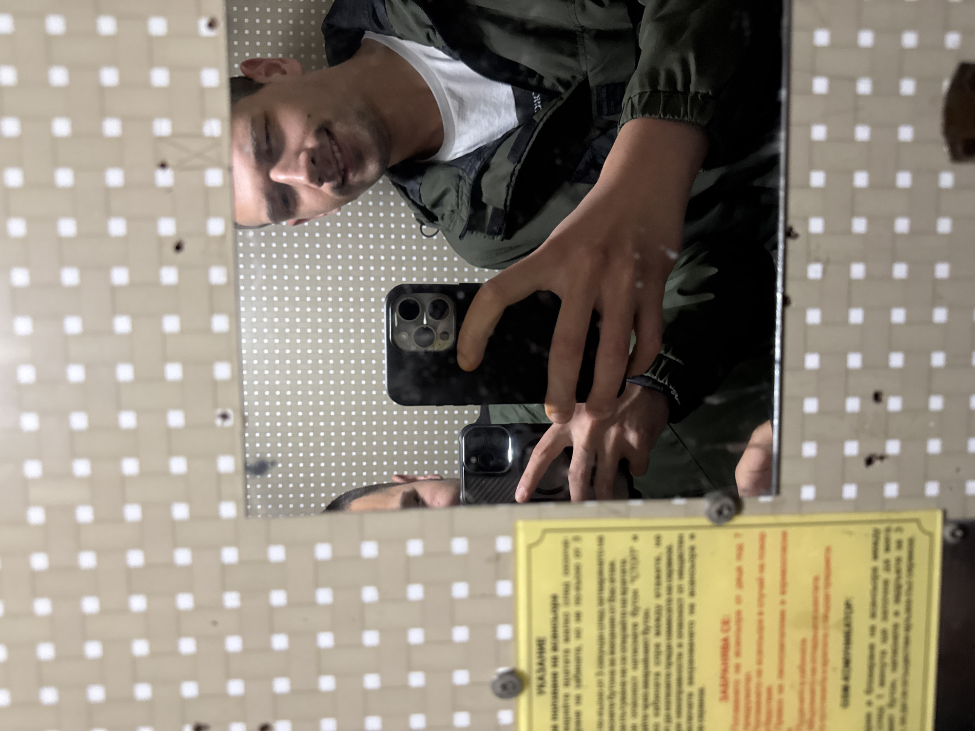 A man is taking a selfie in a mirror. He is wearing a white t-shirt and a dark green jacket. The mirror is square and is mounted on a wall with a beige and white checkered pattern. There is a yellow sign with text in a foreign language attached to the lower right corner of the mirror. A second reflection of the man is visible near the sign. The location is Unknown, Unknown.