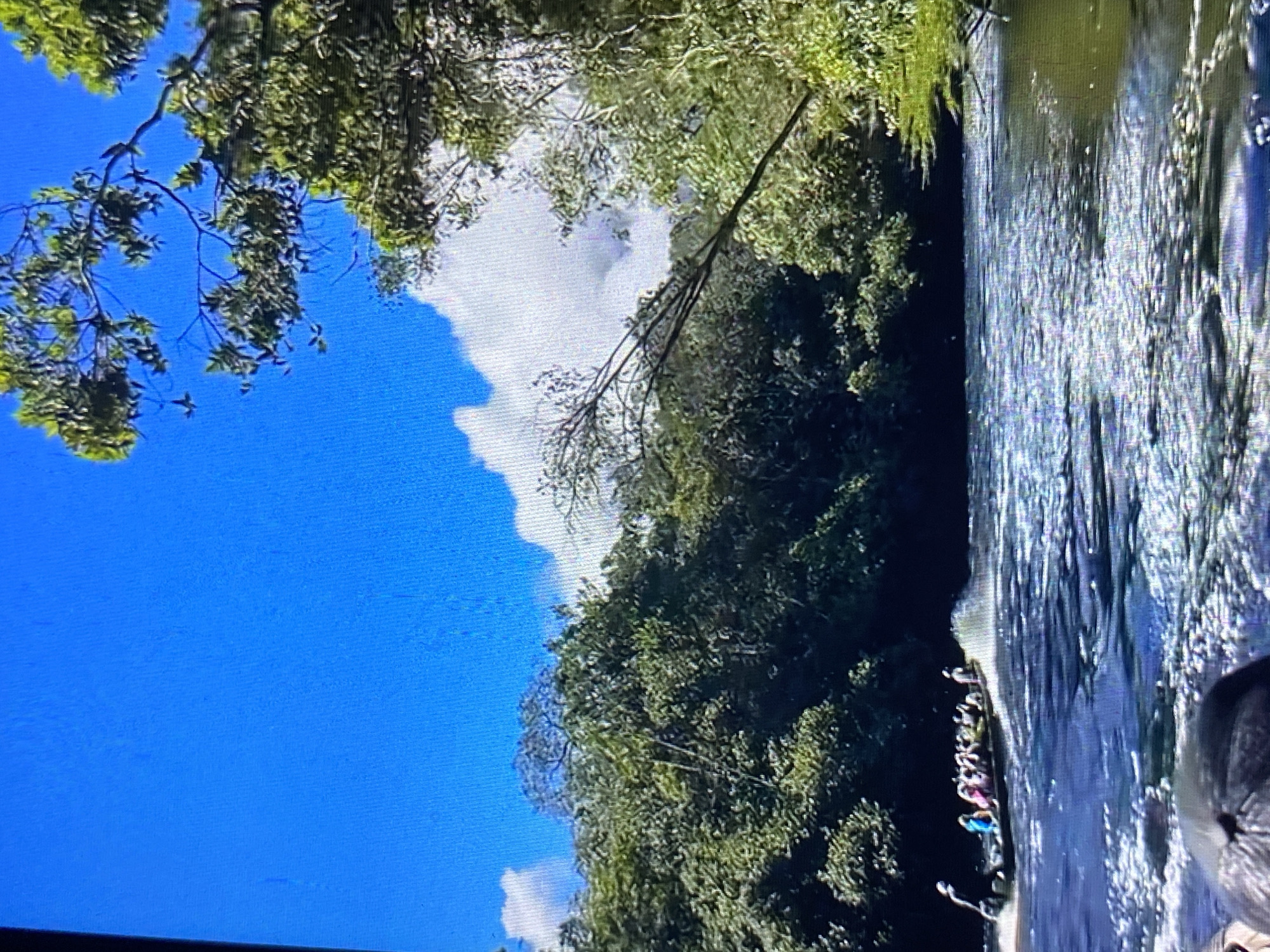 The image shows a waterfall flowing over rocks. A group of people is standing near the base of the waterfall. Lush green vegetation covers the surrounding area. The sky is blue with scattered white clouds. The location is Tinaquillo, Venezuela.
