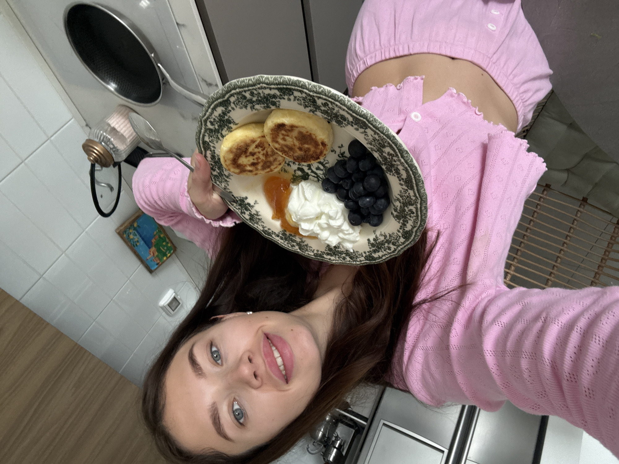 The image is a selfie taken by a young woman in a kitchen or dining area, with the camera oriented counter-clockwise. The woman, with long dark brown hair, blue eyes, and light pink lipstick, smiles warmly at the viewer. She is dressed in a light pink, possibly knit or crocheted, cropped top revealing her midriff, paired with what appears to be a matching button-up cardigan.
She holds a decorative ceramic plate with an intricate dark green floral pattern around its rim. The plate contains a delicious-looking meal, likely breakfast or brunch: two golden-brown, round pancakes or syrniki (cheese fritters), a dollop of white cream, a spoonful of orange-yellow jam or preserves, and a cluster of dark blue grapes. A silver spoon rests on the plate.
In the background, typical kitchen elements are visible, including a silver frying pan with a black non-stick surface, a clear glass jar with a golden lid and a handle (possibly a salt or sugar shaker with a spoon inside), and white tiled walls. A section of light wooden flooring is also seen, along with a small framed picture or artwork on the wall. Part of a wicker or rattan piece of furniture is visible to the right. The woman's happy expression suggests contentment. There is no visible text in the image. While the location context of Tel-Aviv, Israel is provided, the image itself does not contain specific geographical cues.