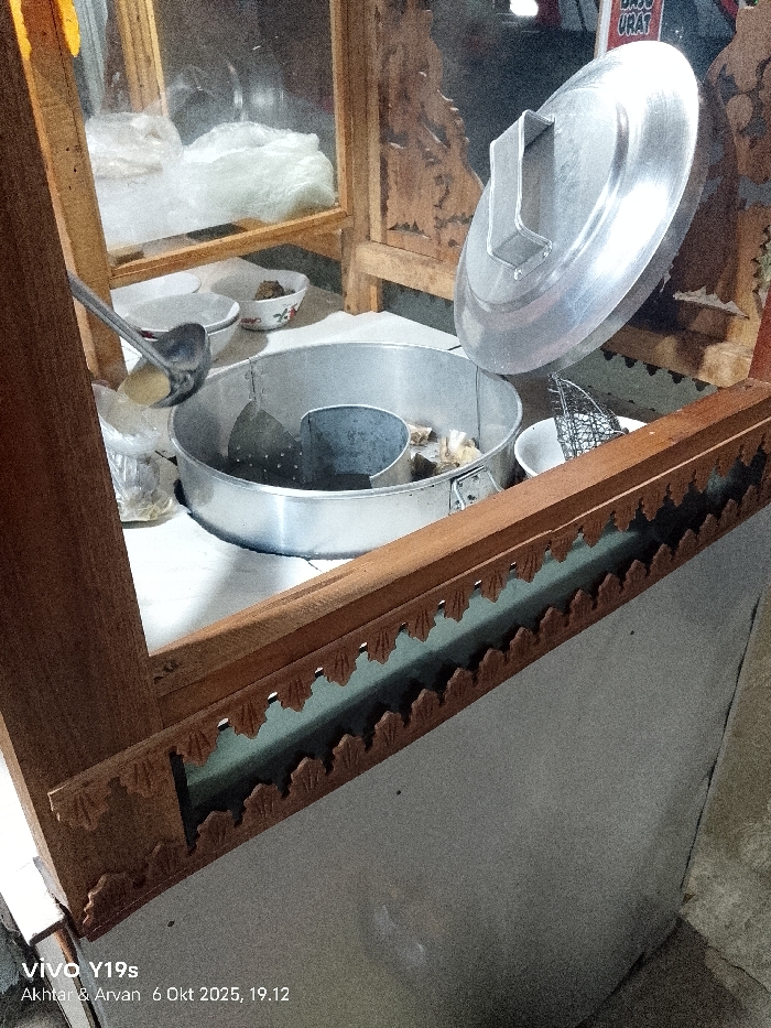 The image displays a food stall setup, likely for serving meatballs, located in Kedungdawa, Indonesia, captured on October 6, 2025, at 19:12. The central object is a large, circular aluminum pot, partially filled with liquid and solid food items, featuring an internal perforated metal divider. A domed aluminum lid is propped open, leaning against the rear of the stall. To the left of the pot, a metal ladle rests within a small white ceramic bowl containing a yellowish liquid, presumably broth. Adjacent to this, another white ceramic bowl holds dark, chunky food items. Behind these, white opaque contents are visible within clear plastic bags. To the right of the main pot, a smaller white ceramic bowl contains a metal wire mesh strainer. The entire counter area is enclosed by a wooden framework, which includes carved decorative details along its front horizontal beam and clear glass panels. A partially visible sign in the blurred background indicates "BASO URAT." No individuals are present in the scene.