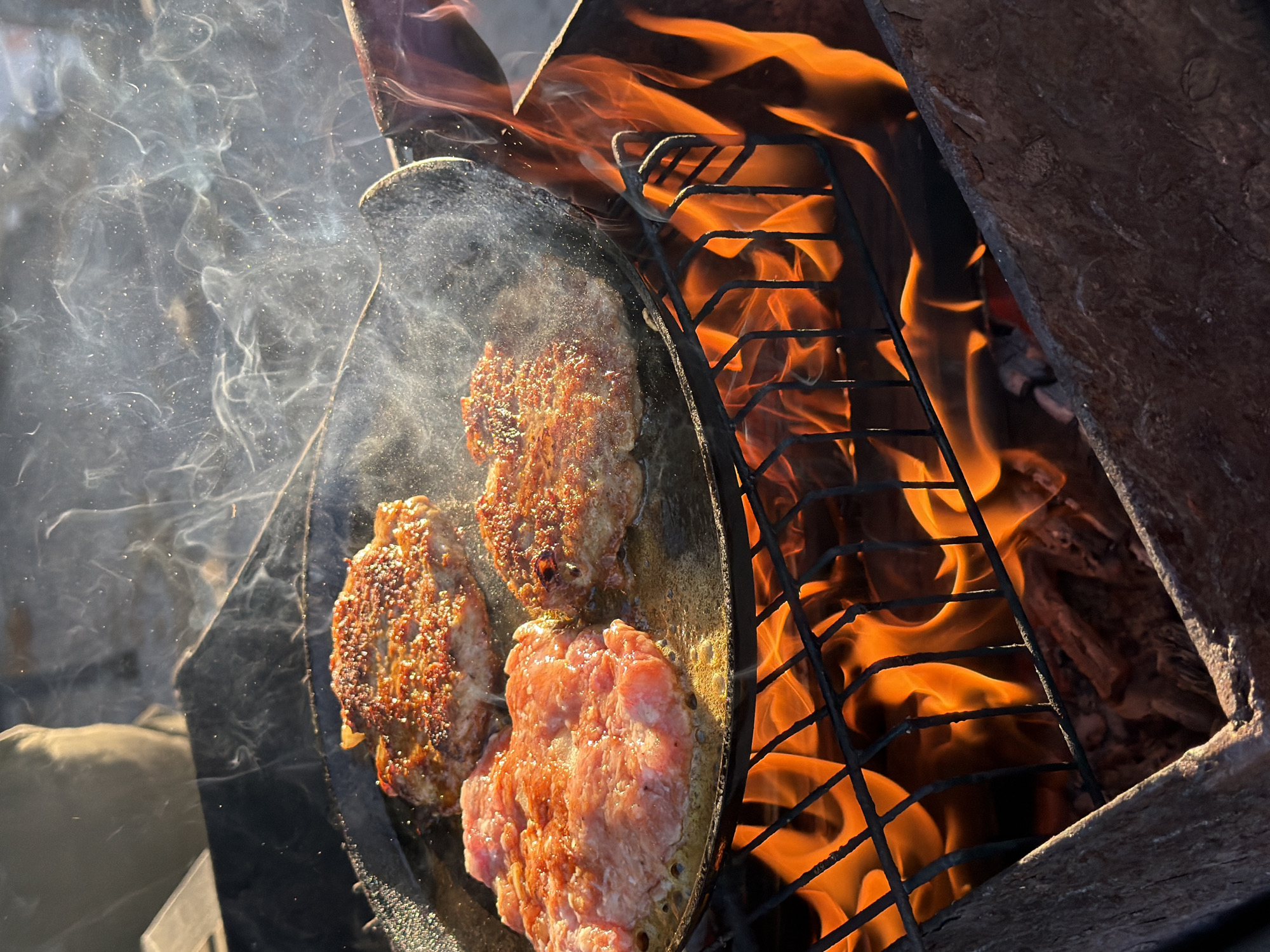 The image depicts three patties cooking in a frying pan over an open fire, emitting smoke. The fire is contained within a structure made of wood and metal grating. The patties are at different stages of cooking, with one appearing raw and the others browned. The setting is outdoors, possibly in Большая Шильна, Russia, based on the provided context. No people are prominently visible. The scene suggests a cooking activity taking place over an open fire.