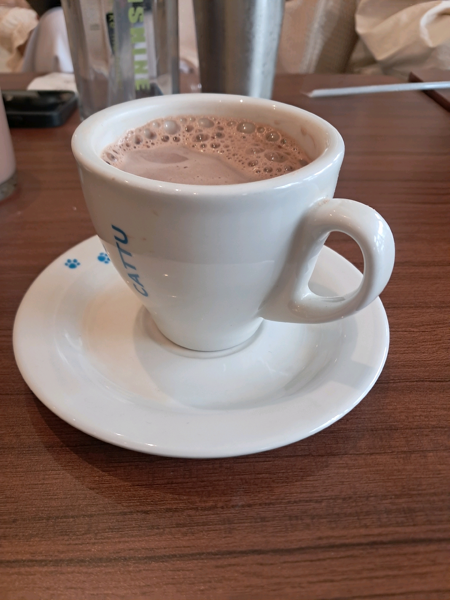 A close-up shot captures a white ceramic cup filled with chocolate milk, topped with a frothy layer, resting on a matching saucer. The cup has "CATTU" written in blue lettering on its side, accompanied by two small blue paw prints. The saucer also features two blue paw prints. The cup and saucer sit on a wooden table with a visible wood grain pattern. In the background, out of focus, are a glass container, a metal container, a white paper napkin, and a white straw. The image is taken from a high angle, with the lighting suggesting an indoor setting. No people are visible in the image. The location in Singkawang, Indonesia is not determinable from the image itself.