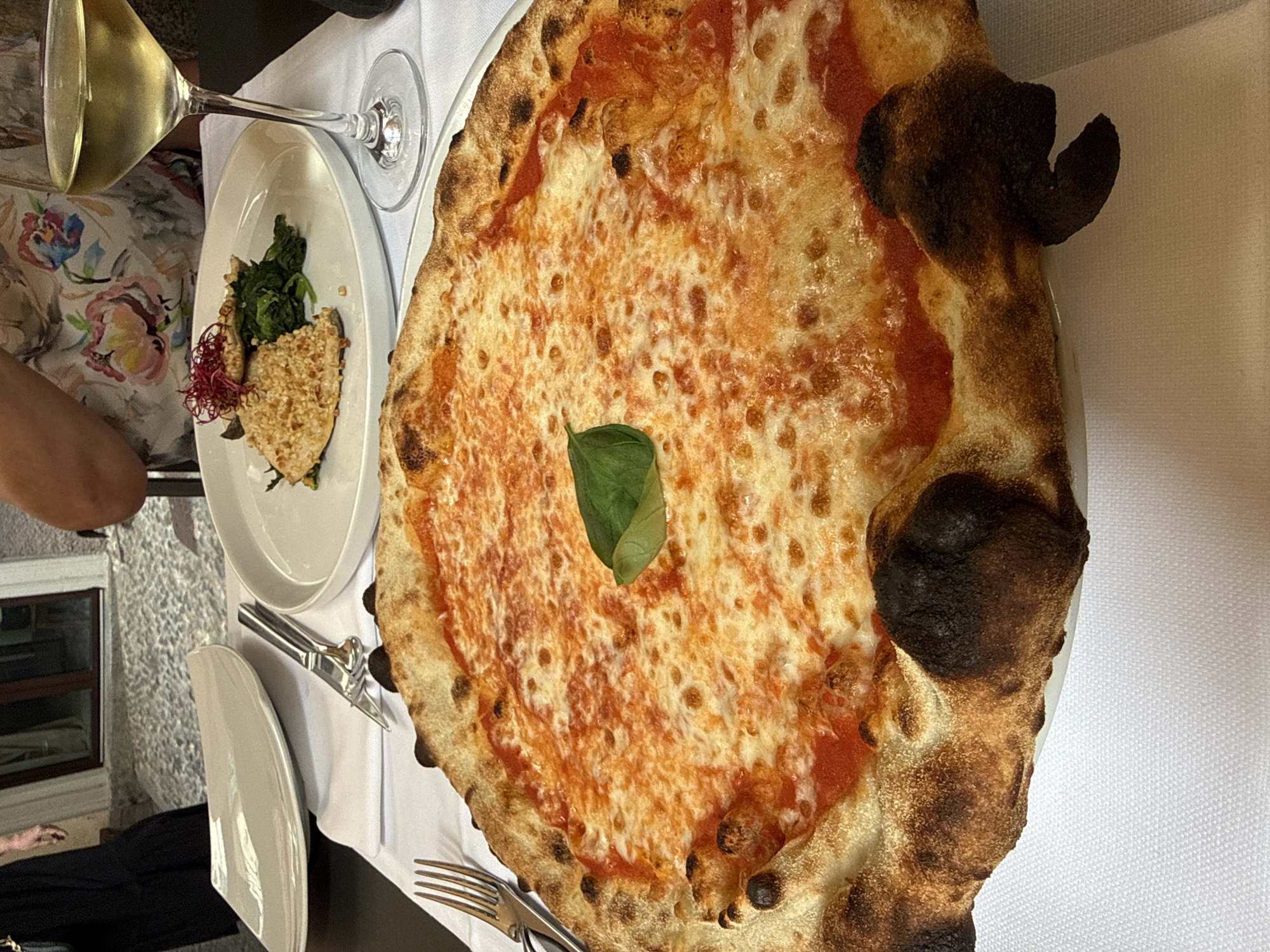 An overhead shot captures a close-up view of a pizza on a white tablecloth. The pizza is circular and topped with tomato sauce and melted mozzarella cheese, with a single green basil leaf placed in the center. The crust is slightly charred in places. To the right of the pizza is a white plate with a serving of food, including what appears to be a piece of fish coated in nuts, served over a bed of spinach, and garnished with red threads. A wine glass filled with white wine sits beside the plate. In the background, a person wearing a floral-patterned top can be seen. The setting appears to be an outdoor dining area, with cobblestone pavement visible behind the table. The scene suggests a meal at a restaurant in San Giovanni, Italy, given the distinct architectural elements suggestive of Italian regions and the presence of food and drink commonly found in Italian cuisine.