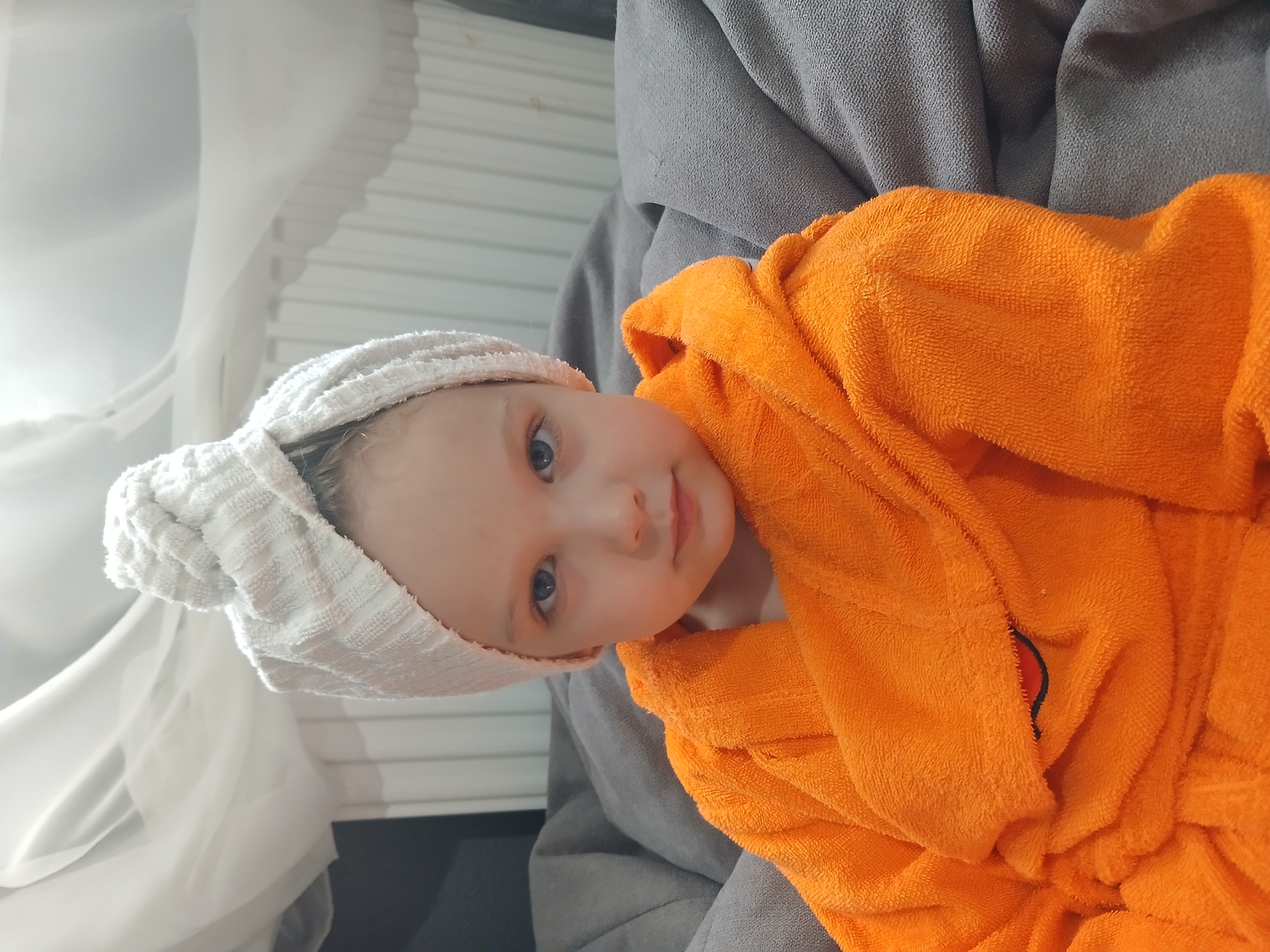 A young child, appearing to be female, sits on a grey upholstered surface, possibly a beanbag chair or sofa. The child is wearing an orange terrycloth bathrobe and a white terrycloth towel turban on her head. Her eyes are open and she is looking directly at the camera. The background shows a white sheer curtain and a radiator. The image is likely taken indoors in a residential setting. There is no visible indication of the specific location within Yekaterinburg, Russia.